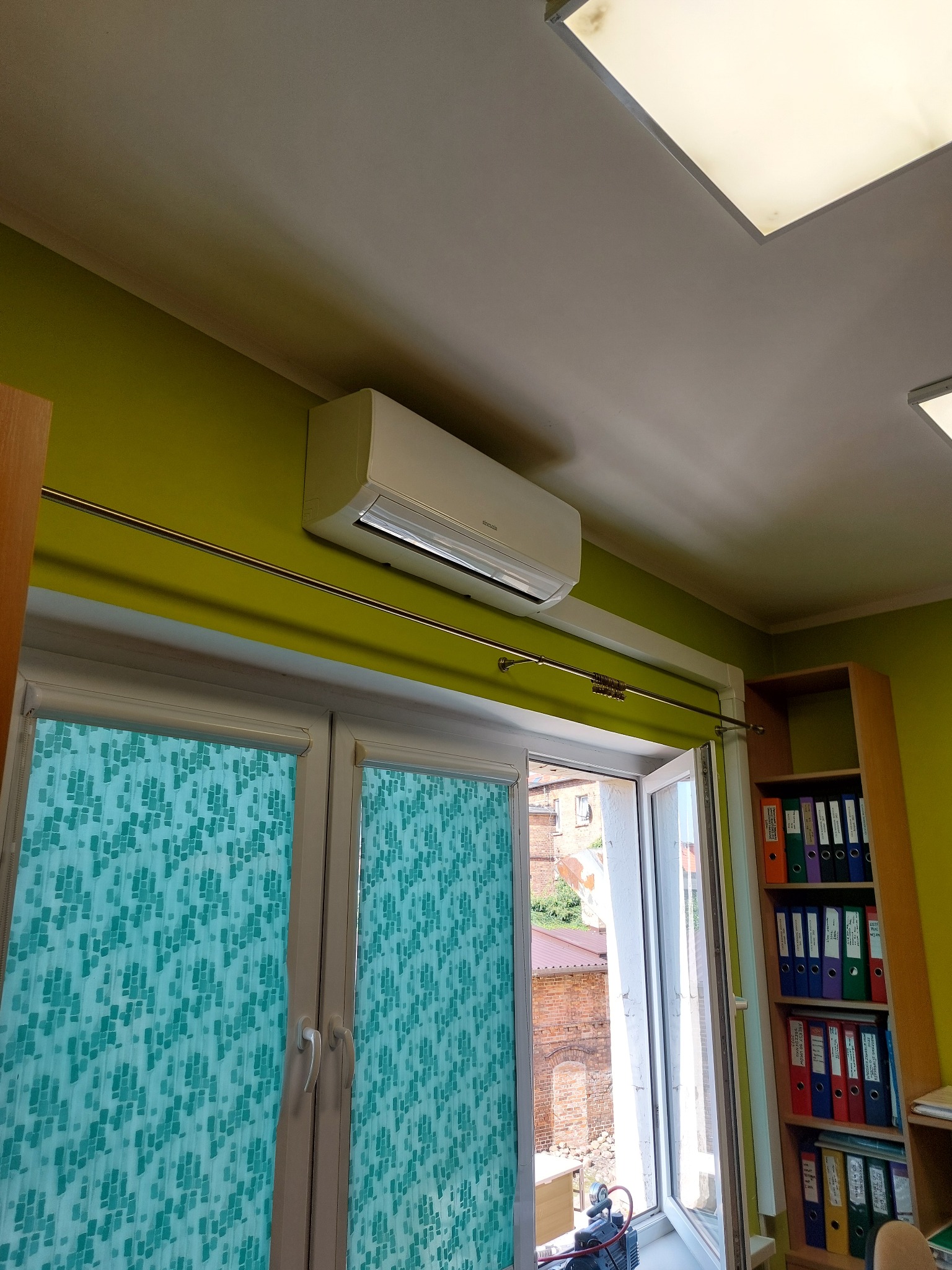 Biała jednostka wewnętrzna klimatyzatora zamontowana nad oknem z turkusowymi roletami w pomieszczeniu biurowym z zielonymi ścianami i regałem z segregatorami.