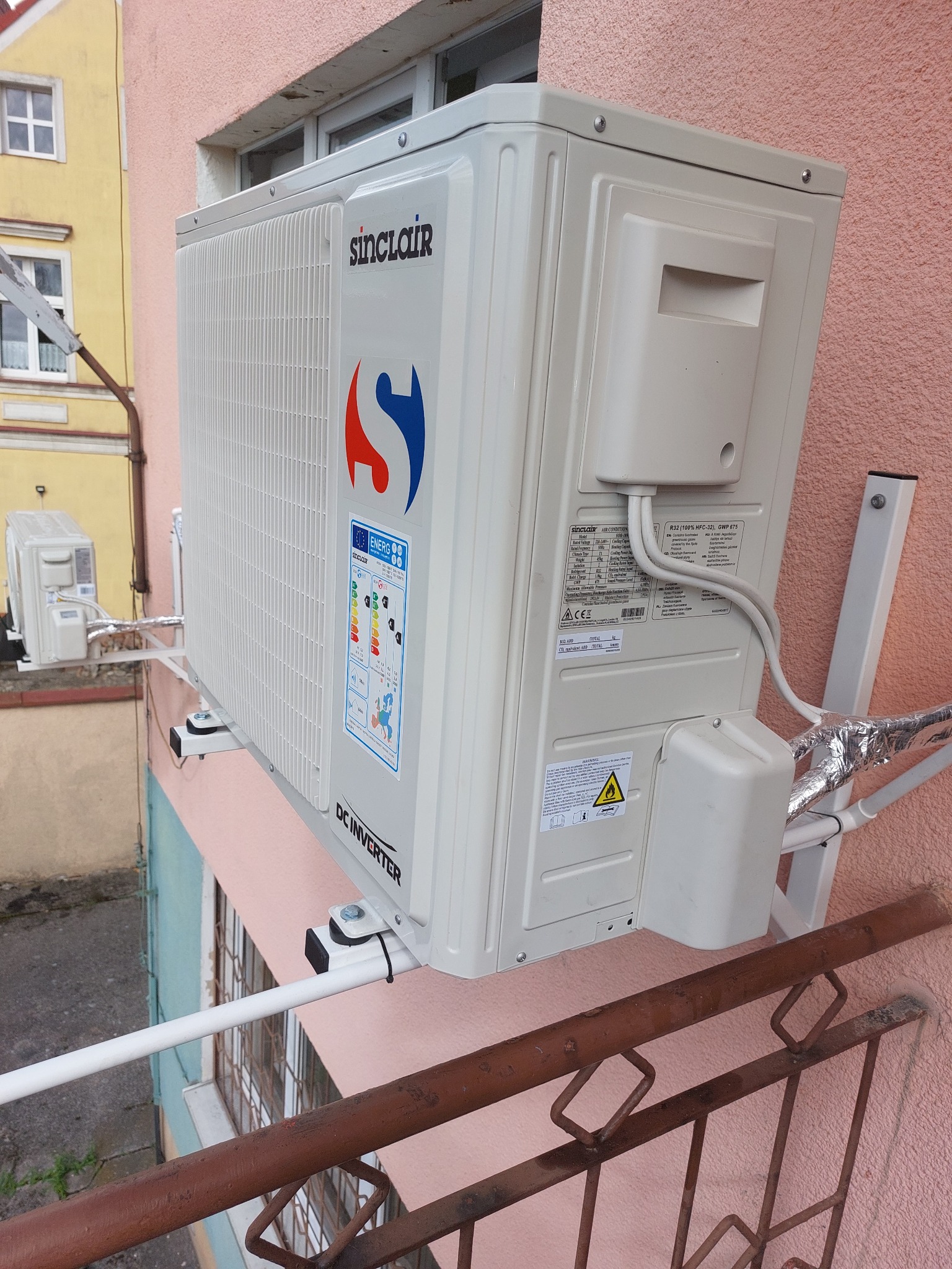 Zewnętrzna jednostka klimatyzacji Sinclair zamontowana na ścianie budynku, widoczna z boku, z etykietą energetyczną i przewodami chłodniczymi owiniętymi folią aluminiową, z metalową balustradą...