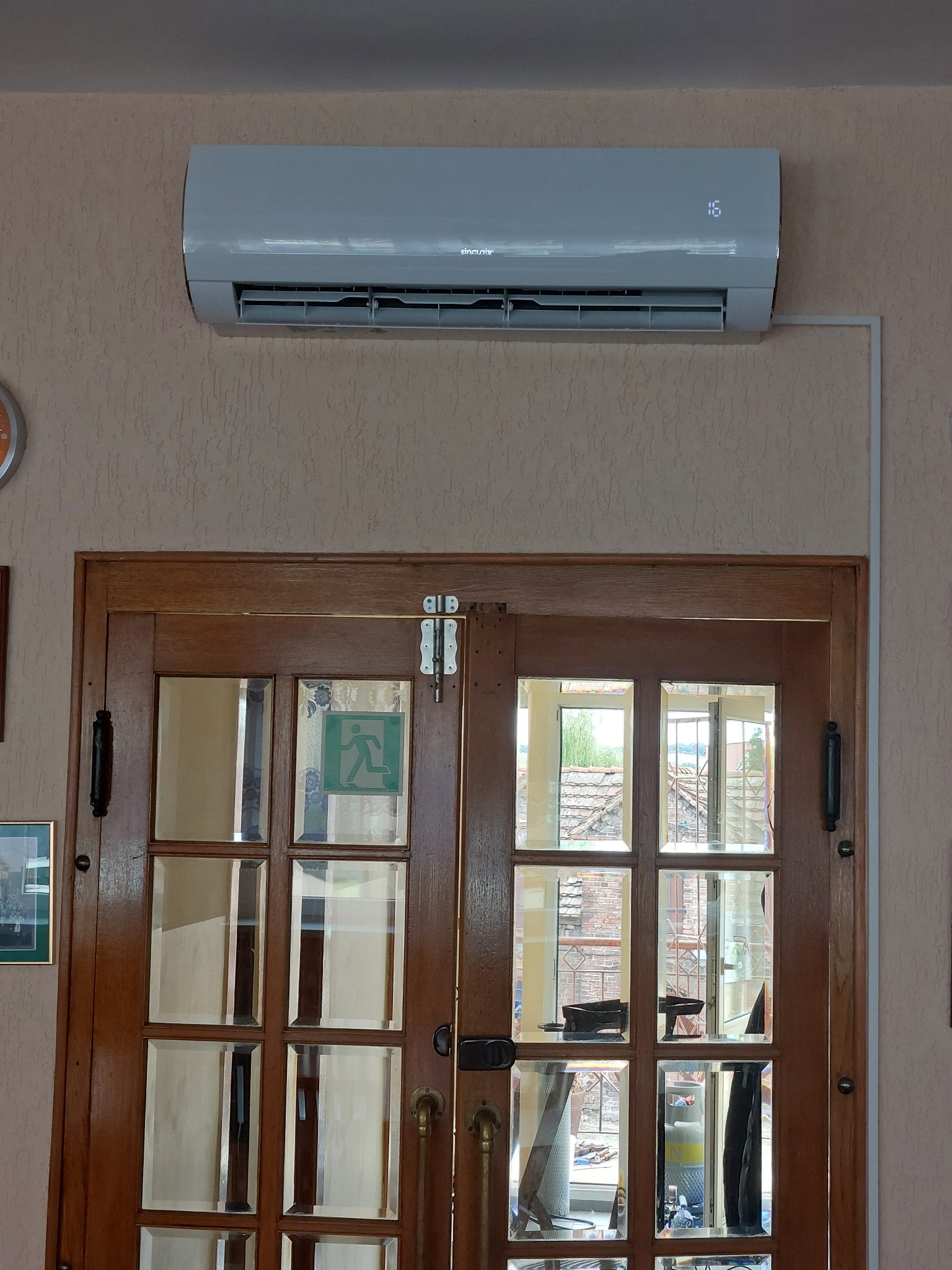 Szara jednostka klimatyzacji zamontowana nad drewnianymi drzwiami z szybami, widoczne okablowanie i wyświetlacz temperatury.