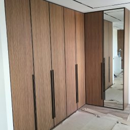 VS FURNITURE SPÓŁKA Z OGRANICZONĄ ODPOWIEDZIALNOŚCIĄ - Wnętrze z narożną zabudową szafy z drewnopodobnymi frontami i czarnymi, długimi uchwytami, uzupełnione lustrem w cienkiej ramie, podłoga zabezpieczona kartonem.