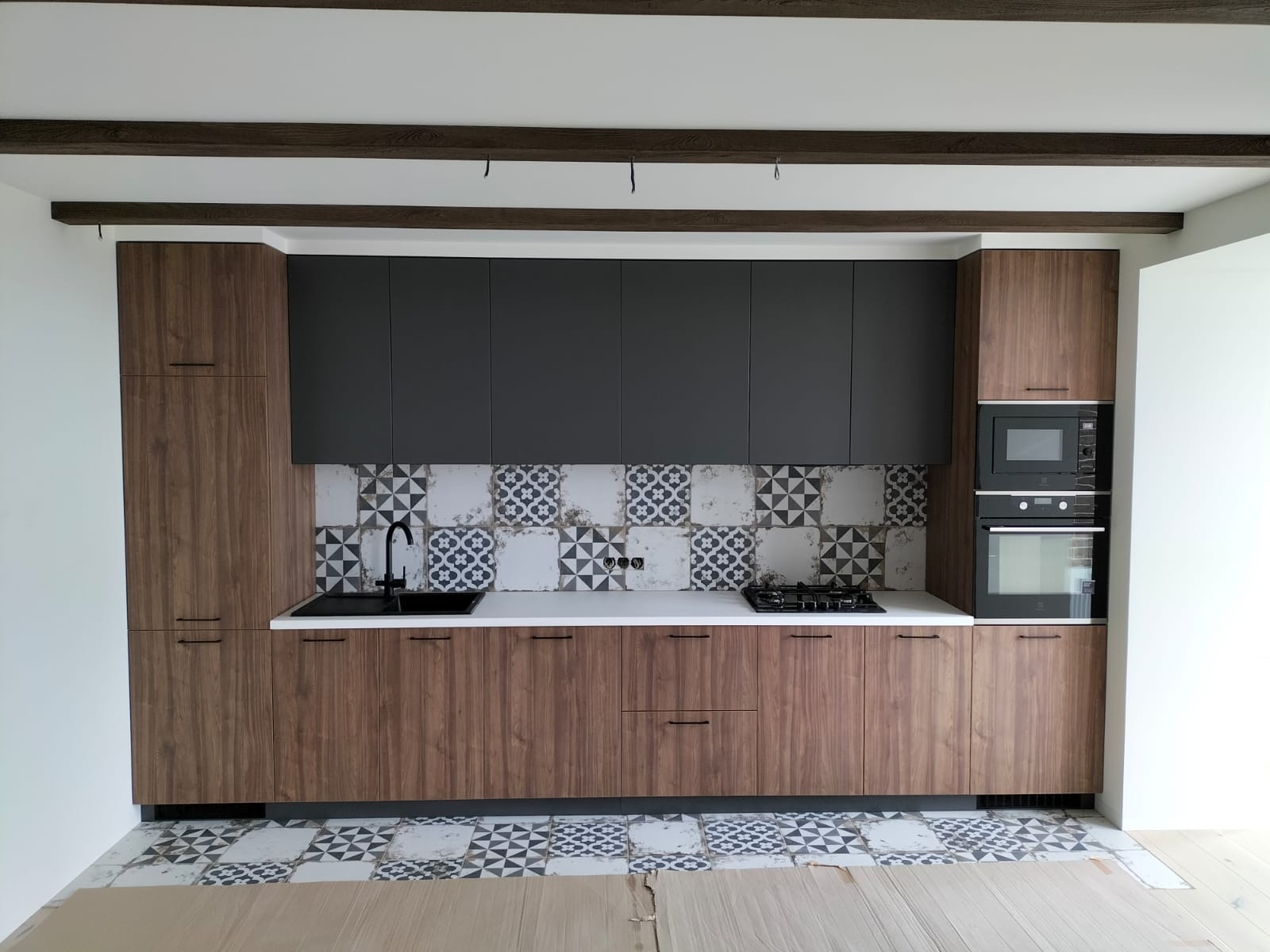 Nowoczesna kuchnia z drewnianymi szafkami dolnymi i grafitowymi górnymi, biały blat, czarna armatura i dekoracyjne płytki na ścianie oraz podłodze z widocznymi belkami stropowymi.