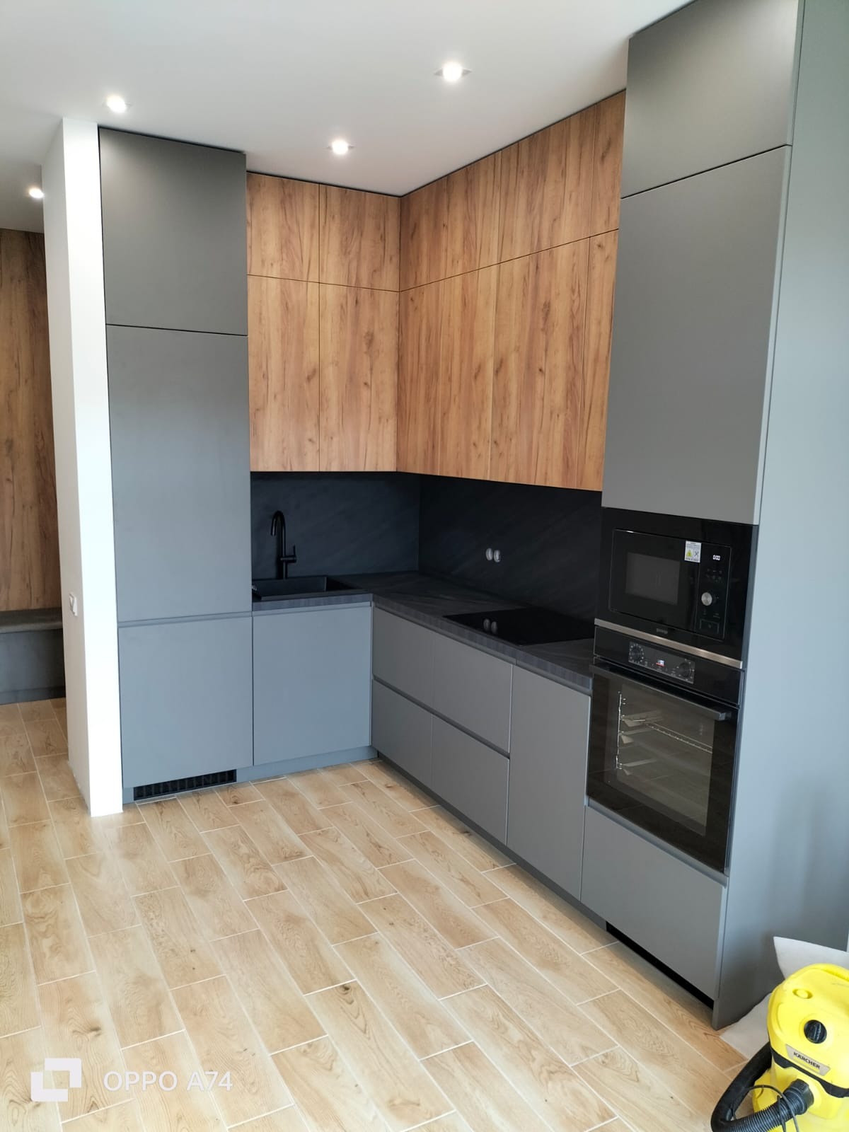 Nowoczesna kuchnia z szarymi szafkami dolnymi i drewnopodobnymi górnymi, czarny zlew i płyta, sprzęt AGD w zabudowie, jasna podłoga imitująca drewno.