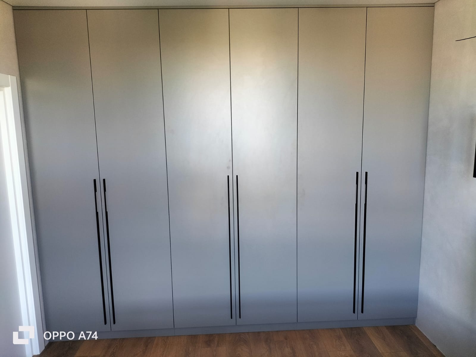 Szafa wnękowa z szarymi frontami i długimi, czarnymi uchwytami, widoczna na tle drewnianej podłogi.