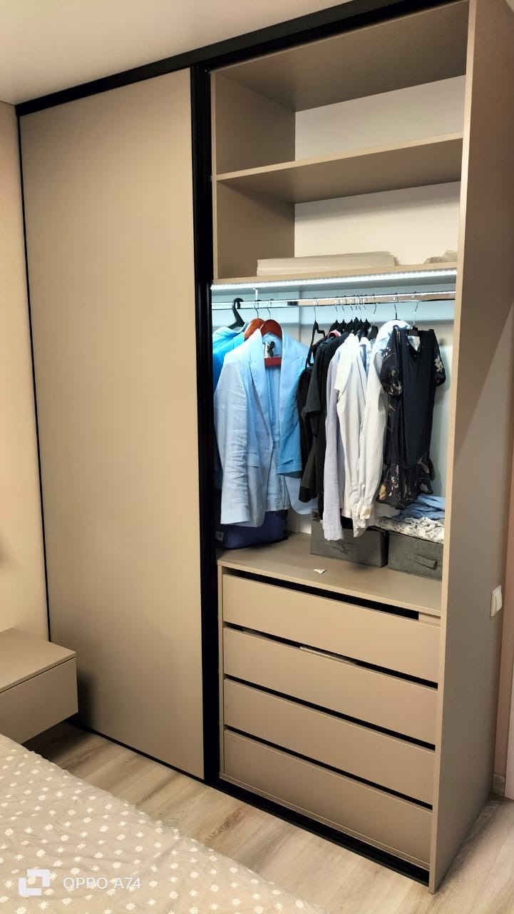 Beżowa szafa przesuwna z czarnymi obramowaniami, częściowo otwarta, ukazująca ubrania na wieszakach, półki i szuflady.