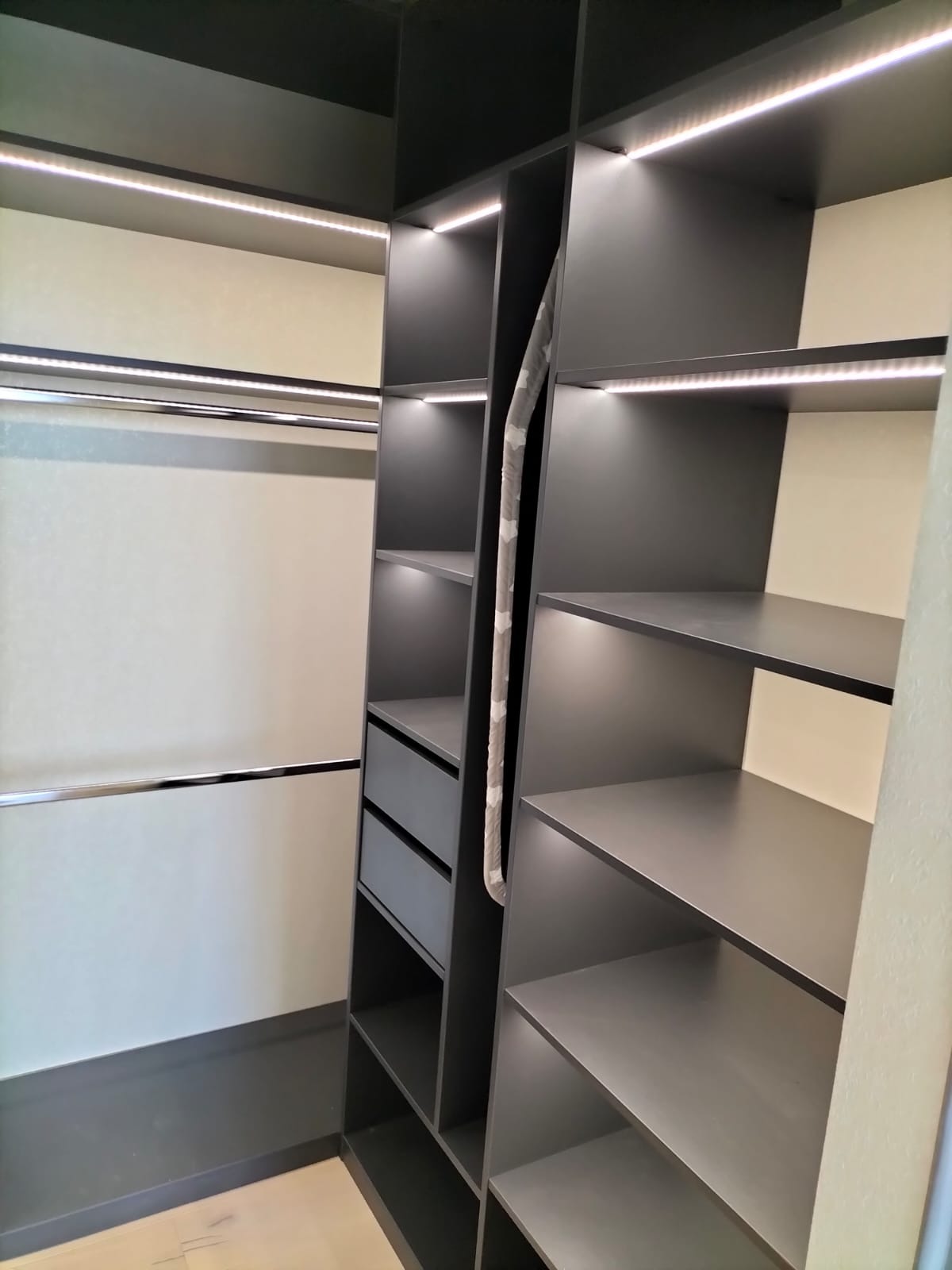 Wnętrze garderoby z szarej zabudowy z półkami, szufladami i drążkami na ubrania, oświetlone listwami LED.