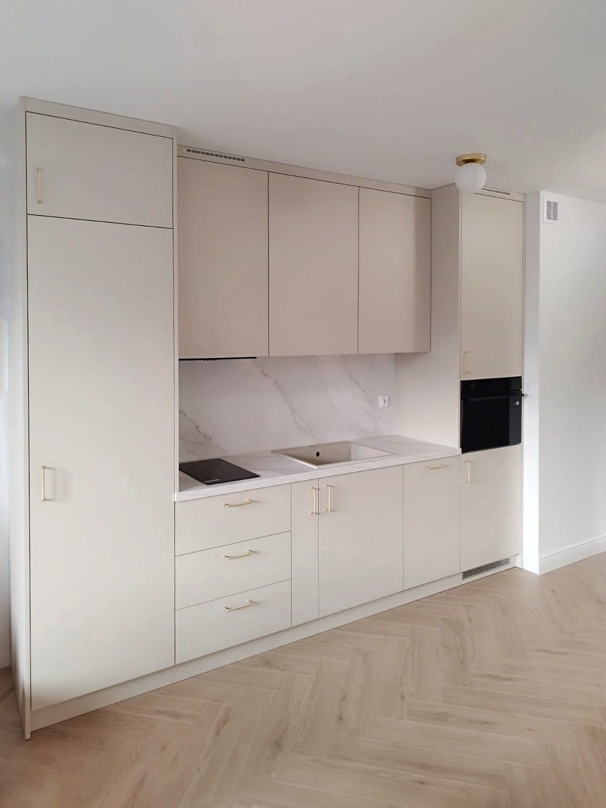 Jasna zabudowa kuchenna z beżowymi frontami i złotymi uchwytami, marmurowy backsplash, płyta indukcyjna i piekarnik w wysokiej zabudowie. Parkiet w jodełkę na podłodze.
