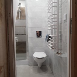 Styl-Dom - Nowoczesna łazienka z szarymi płytkami, wiszącą toaletą, grzejnikiem drabinkowym i kabiną prysznicową z widoczną słuchawką. Na grzejniku powieszony jest ręcznik w odcieniach niebieskiego i szarego.