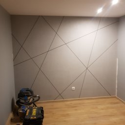 Styl-Dom - Ściana w pokoju pomalowana na szaro z geometrycznym wzorem z ciemnoszarych linii, na podłodze leżą narzędzia budowlane i odkurzacz przemysłowy.