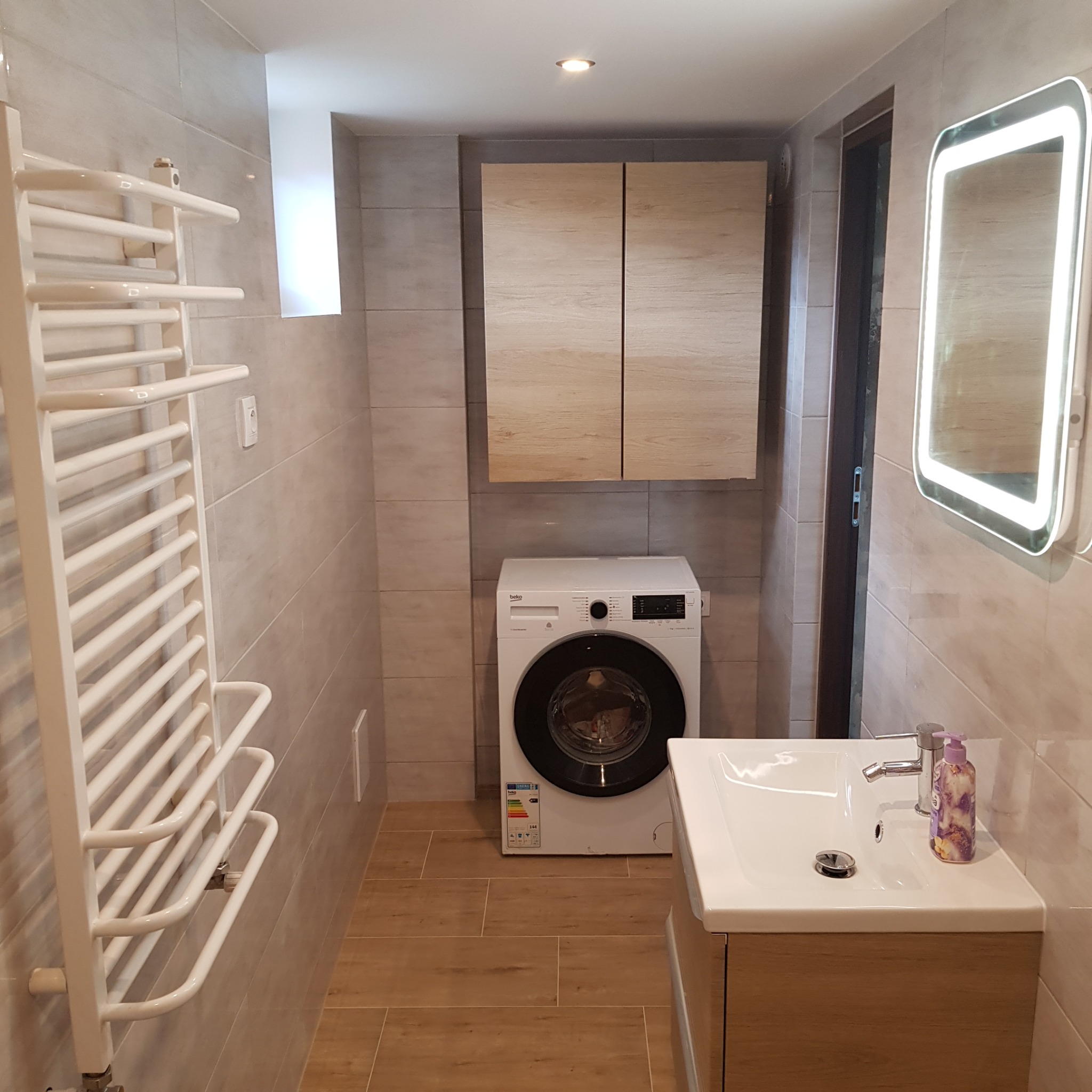 Nowoczesna łazienka z beżowymi płytkami, grzejnikiem drabinkowym, szafką z umywalką, pralką i szafkami wiszącymi, oświetlona lustrem z LED