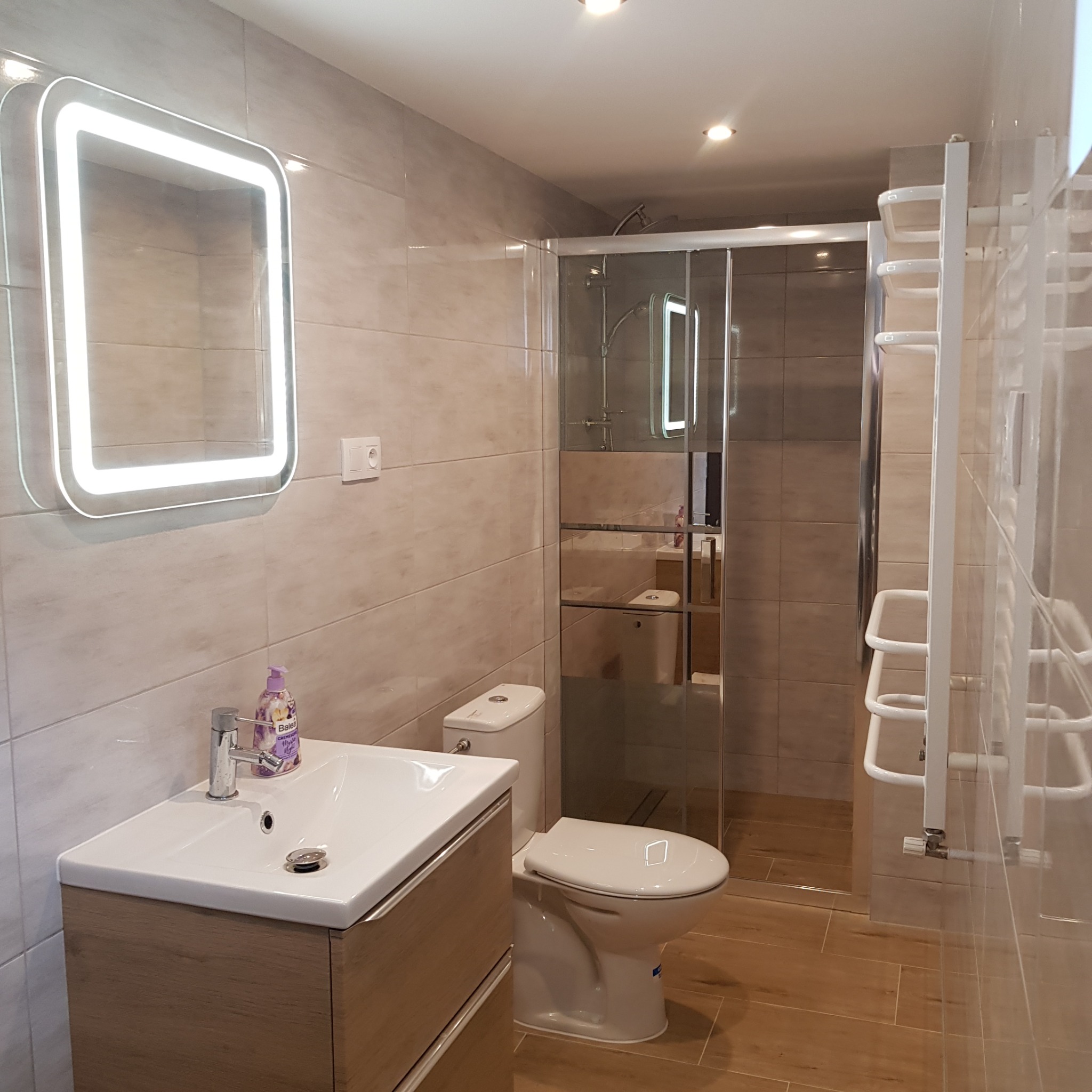 Nowoczesna łazienka z beżowymi płytkami, podświetlanym lustrem, kabiną prysznicową ze szklanymi drzwiami, umywalką z szafką z drewnopodobnym fornirem i białym grzejnikiem drabinkowym.