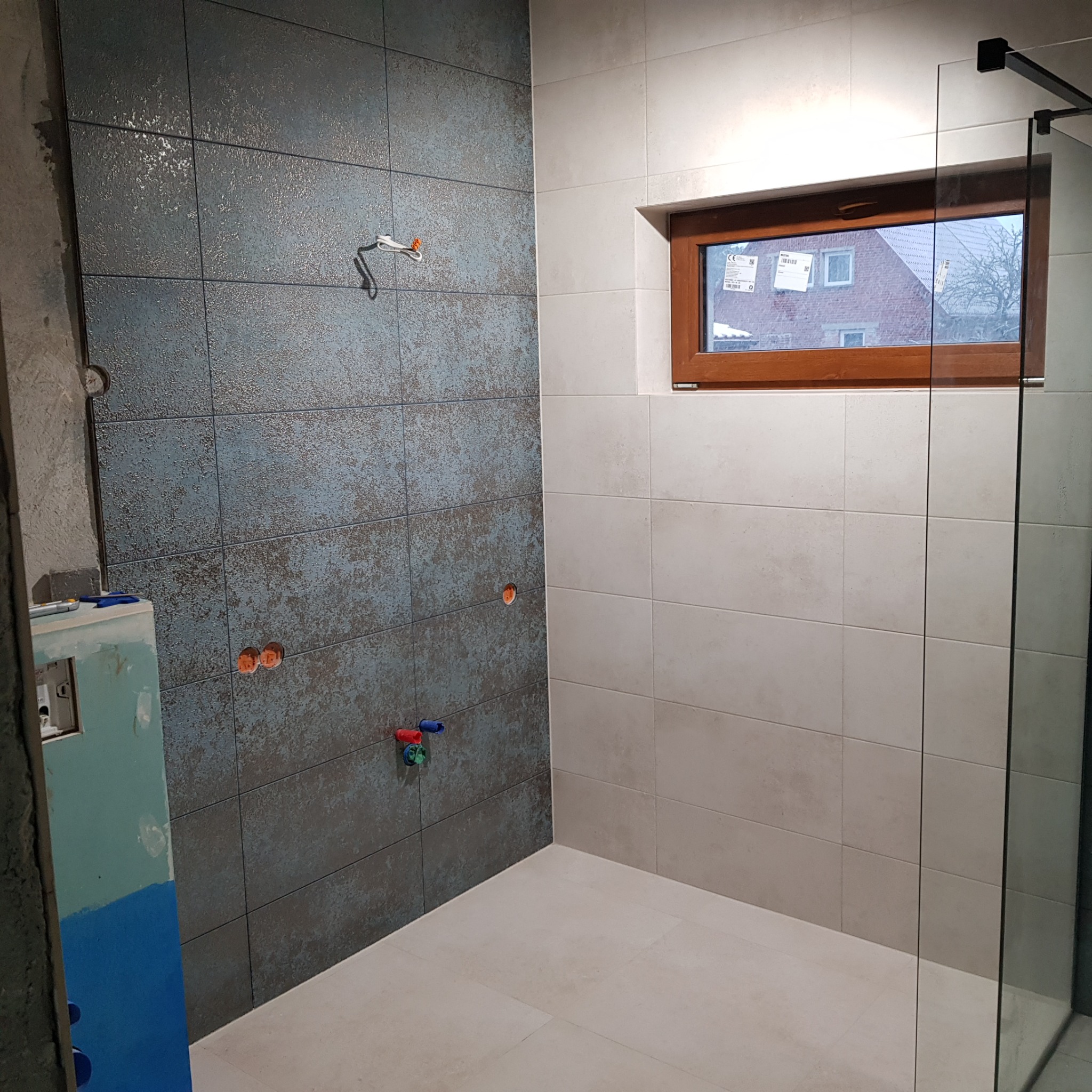 Wnętrze łazienki w trakcie wykańczania: ściana z niebiesko-szarych płytek o nieregularnej fakturze kontrastuje z jasnymi płytkami, widoczne wystające rury instalacji wodnej, okno z drewnianą ramą...