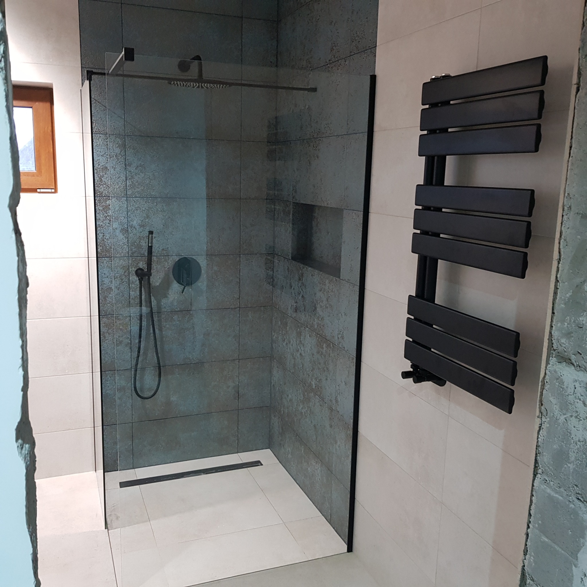 Nowoczesna kabina prysznicowa walk-in z czarnymi elementami: deszczownica, bateria, odpływ liniowy i grzejnik drabinkowy. Ściany wyłożone płytkami w dwóch odcieniach szarości. Fragment ściany...