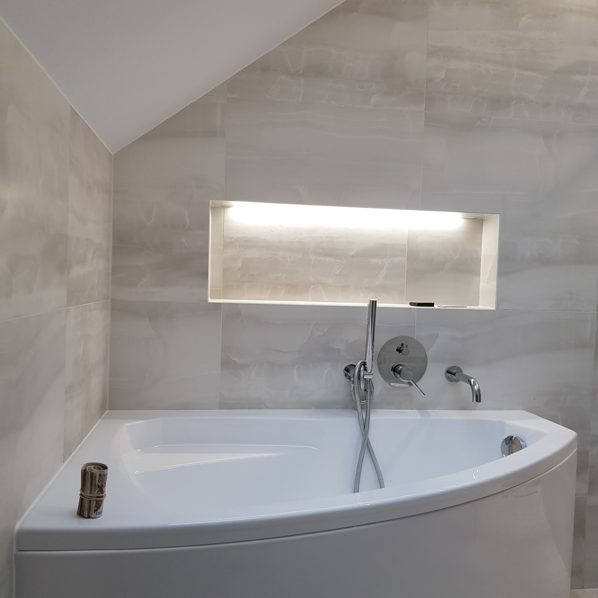 Nowoczesna łazienka z narożną wanną, szarymi płytkami imitującymi marmur i podświetlaną wnęką w ścianie; zwój banknotów leży na krawędzi wanny.