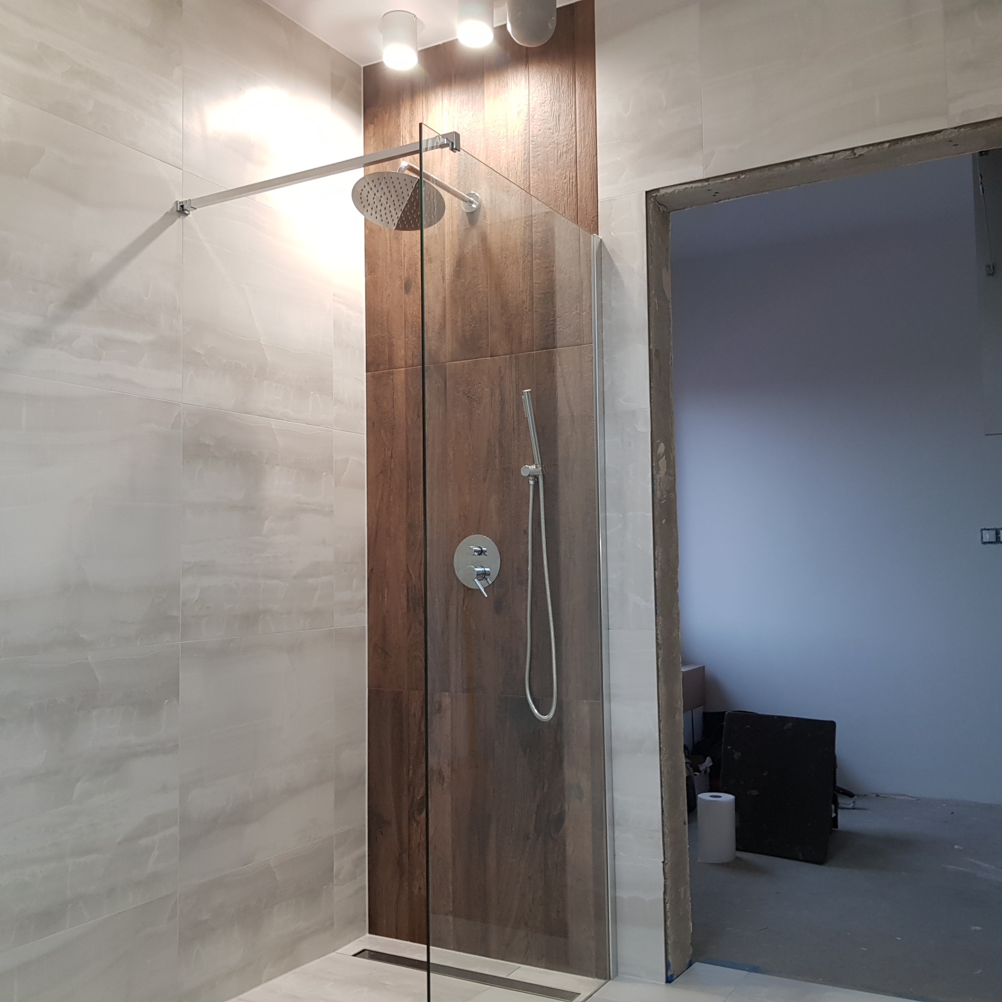 Kabina prysznicowa typu walk-in z deszczownicą, chromowaną armaturą i odpływem liniowym, obłożona płytkami imitującymi marmur i drewno, widoczna na tle surowego otworu drzwiowego.