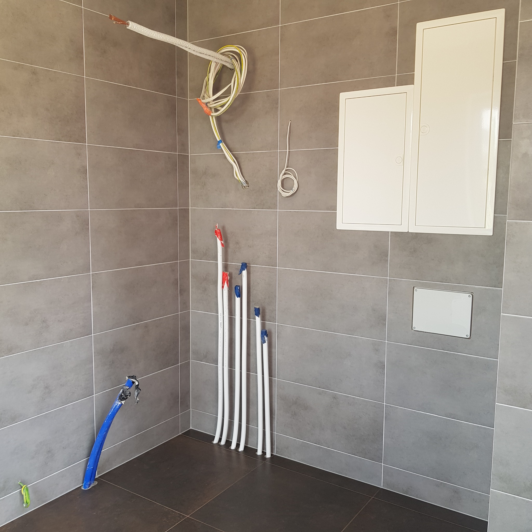 Przygotowania do instalacji w łazience: rury wodne z zaślepkami, wiszące kable elektryczne i dostęp do instalacji ukryty za klapami w szarej glazurze.