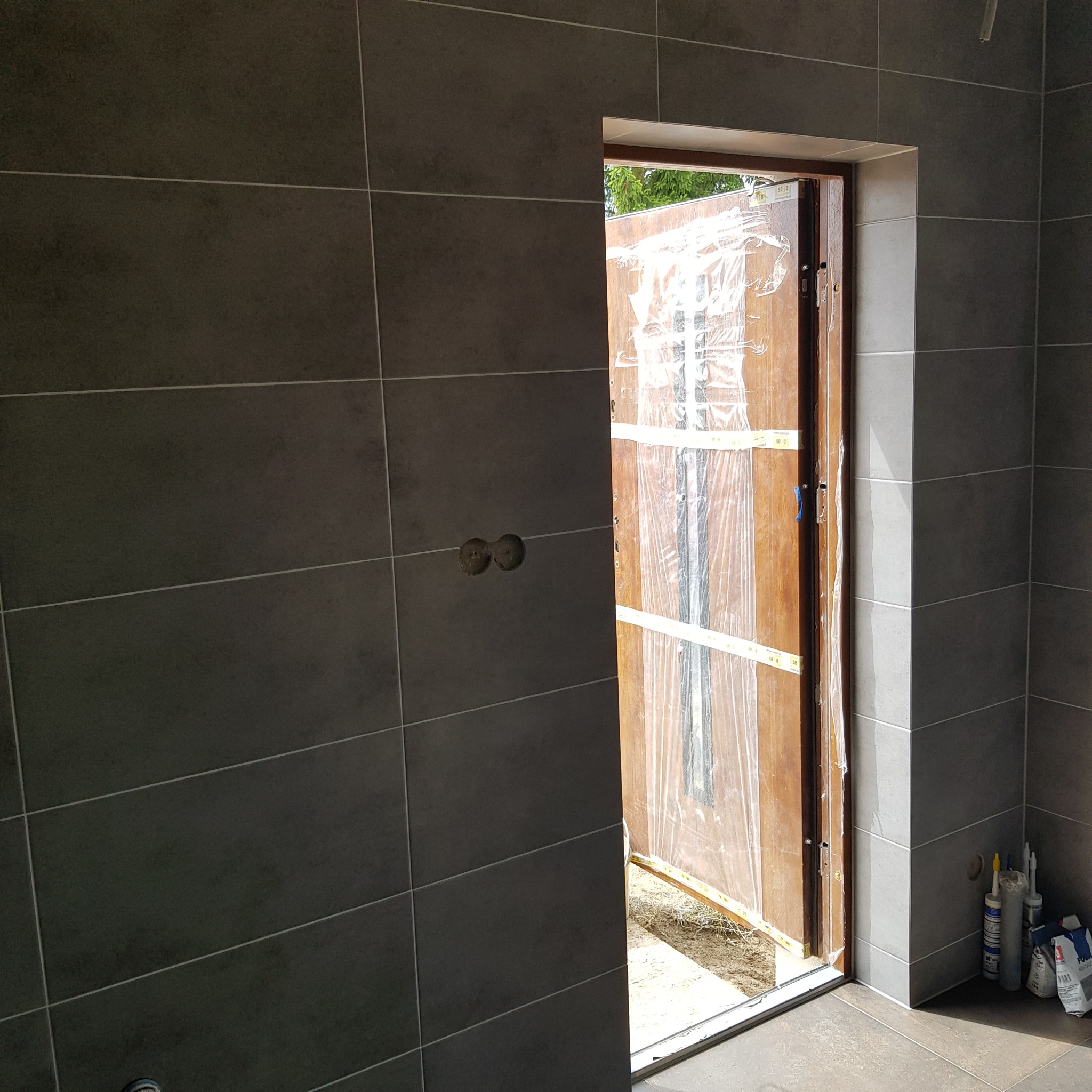 Ściana i podłoga wyłożone ciemnymi płytkami, widoczne puszki instalacyjne i zabezpieczone folią drzwi wejściowe, silikony i zaprawa w workach leżące na podłodze.