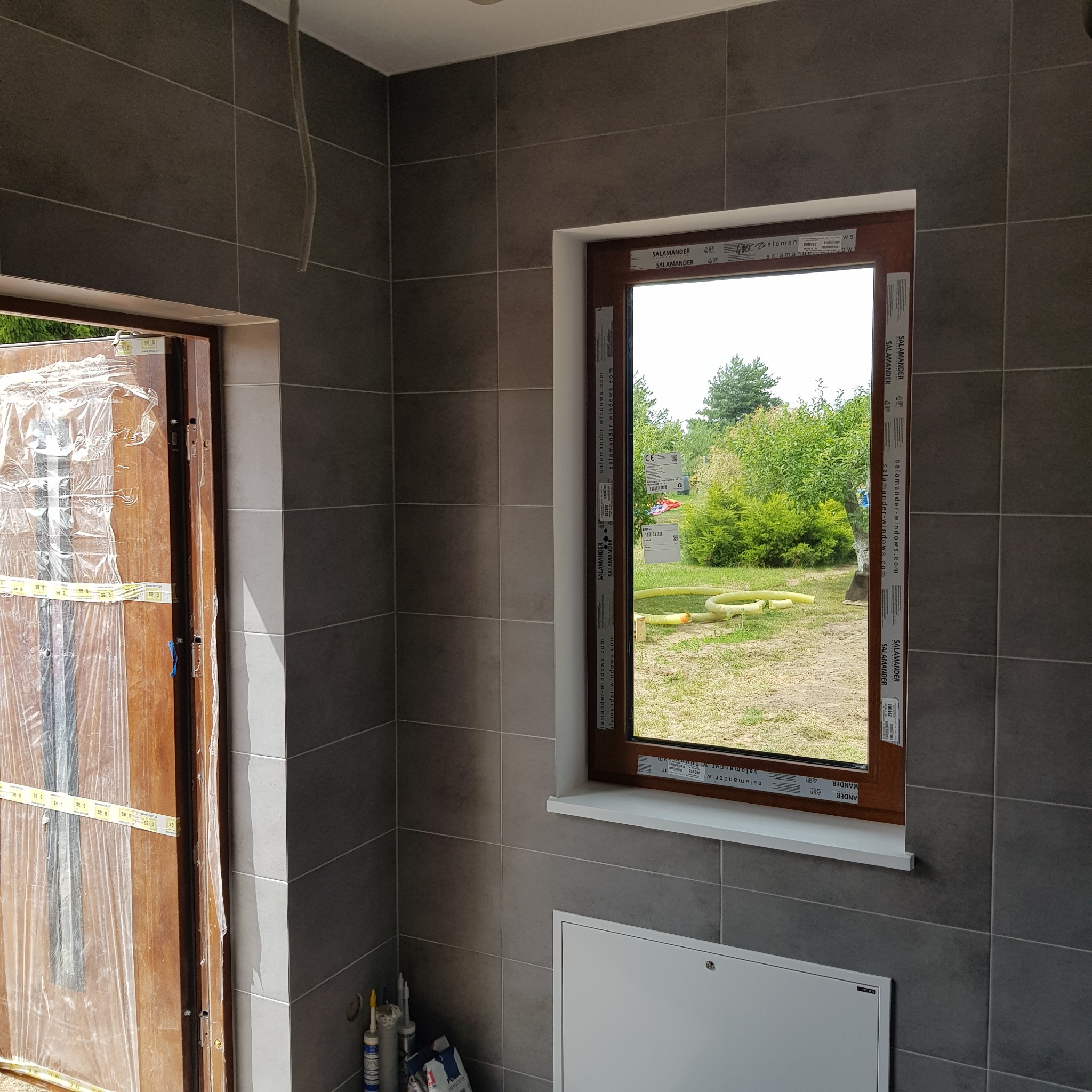 Wykończona łazienka w szarych płytkach, z drewnianą ramą okienną marki Salamander i zabezpieczonym folią wejściem, widoczne silikony i fugi.