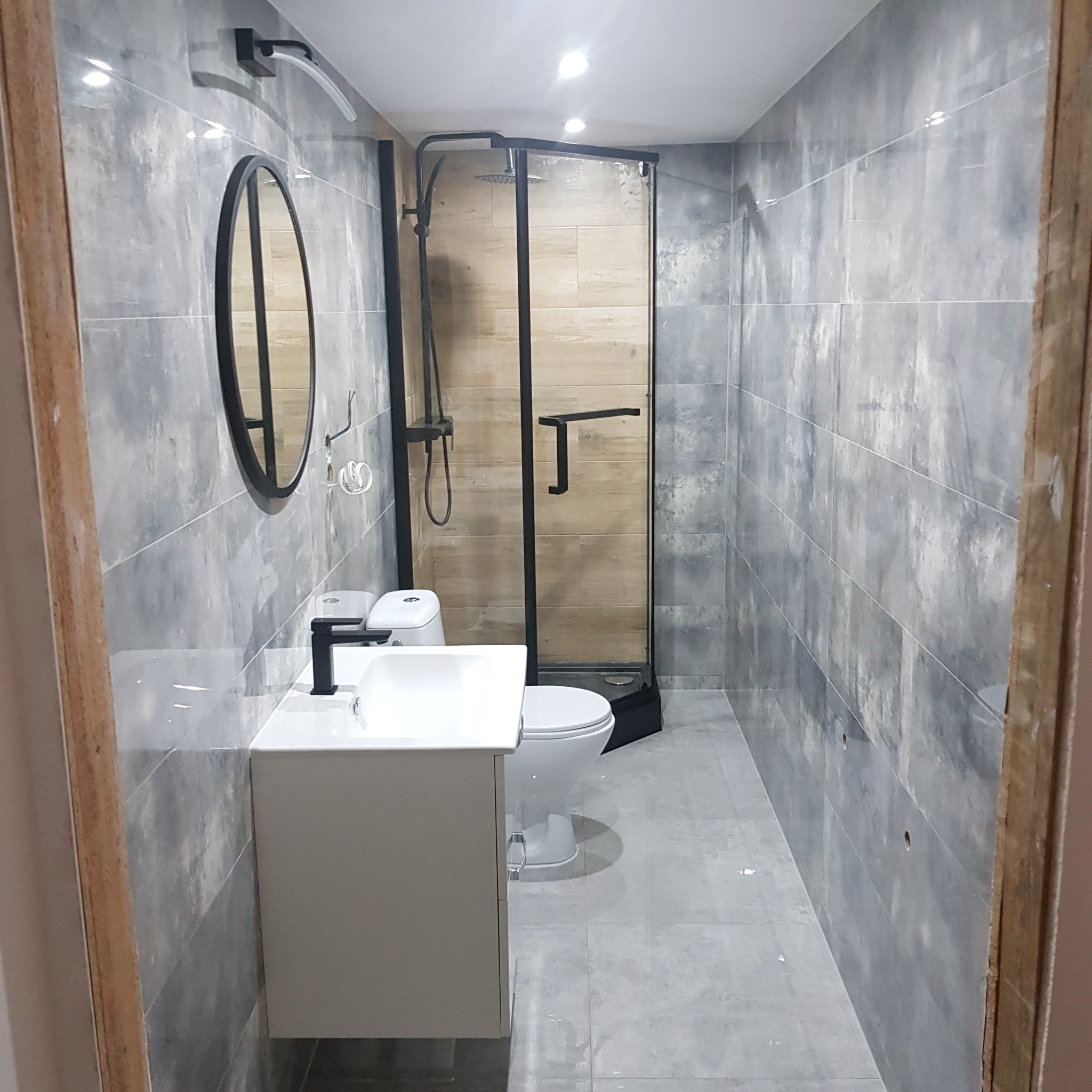 Wykończona łazienka w Choszcznie z szarymi płytkami imitującymi beton, czarną armaturą, owalnym lustrem i kabiną prysznicową z drewnopodobnymi panelami wewnątrz.