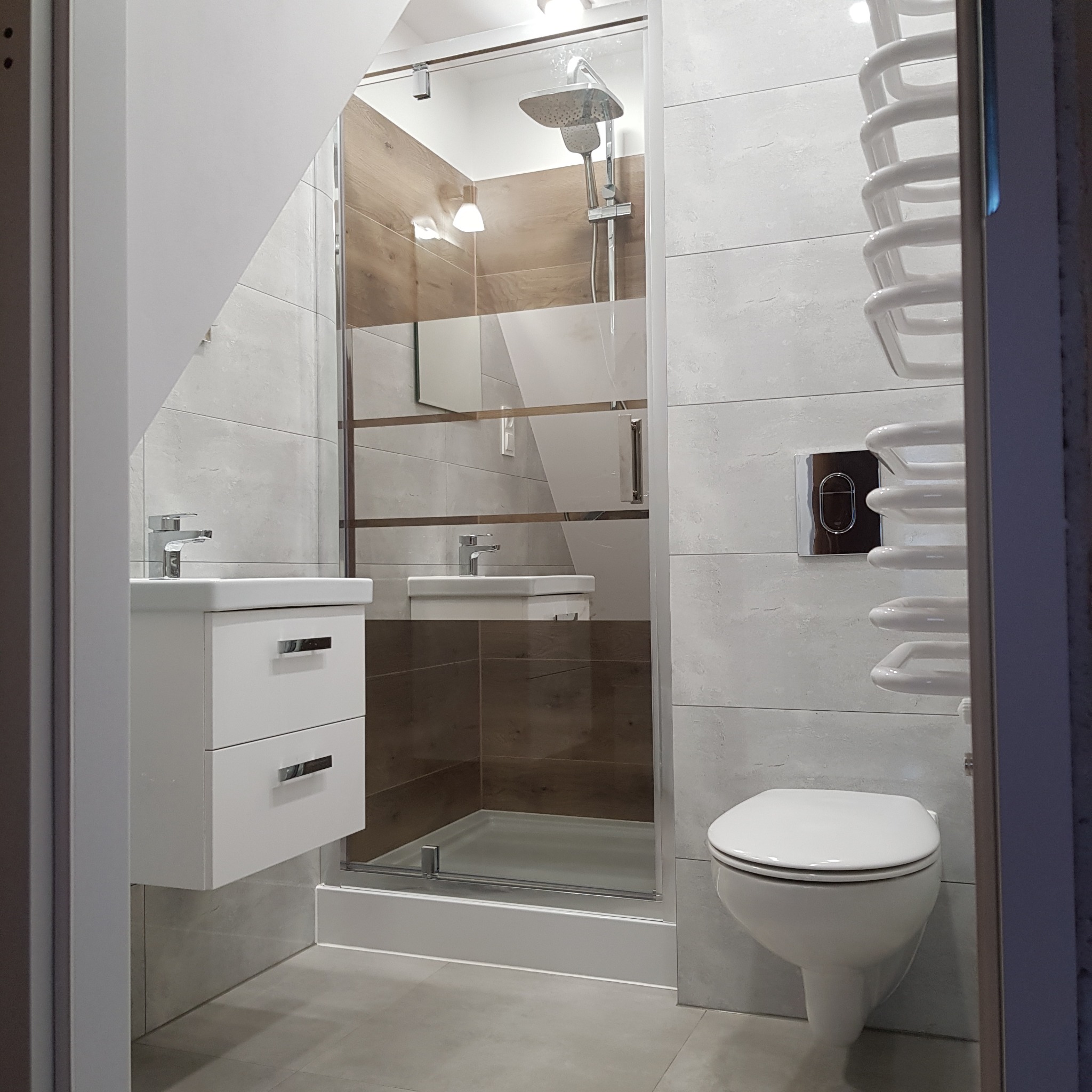 Nowoczesna łazienka z kabiną prysznicową ze szklanymi drzwiami z brązowymi akcentami, umywalką z szafką, wiszącą toaletą i grzejnikiem drabinkowym.