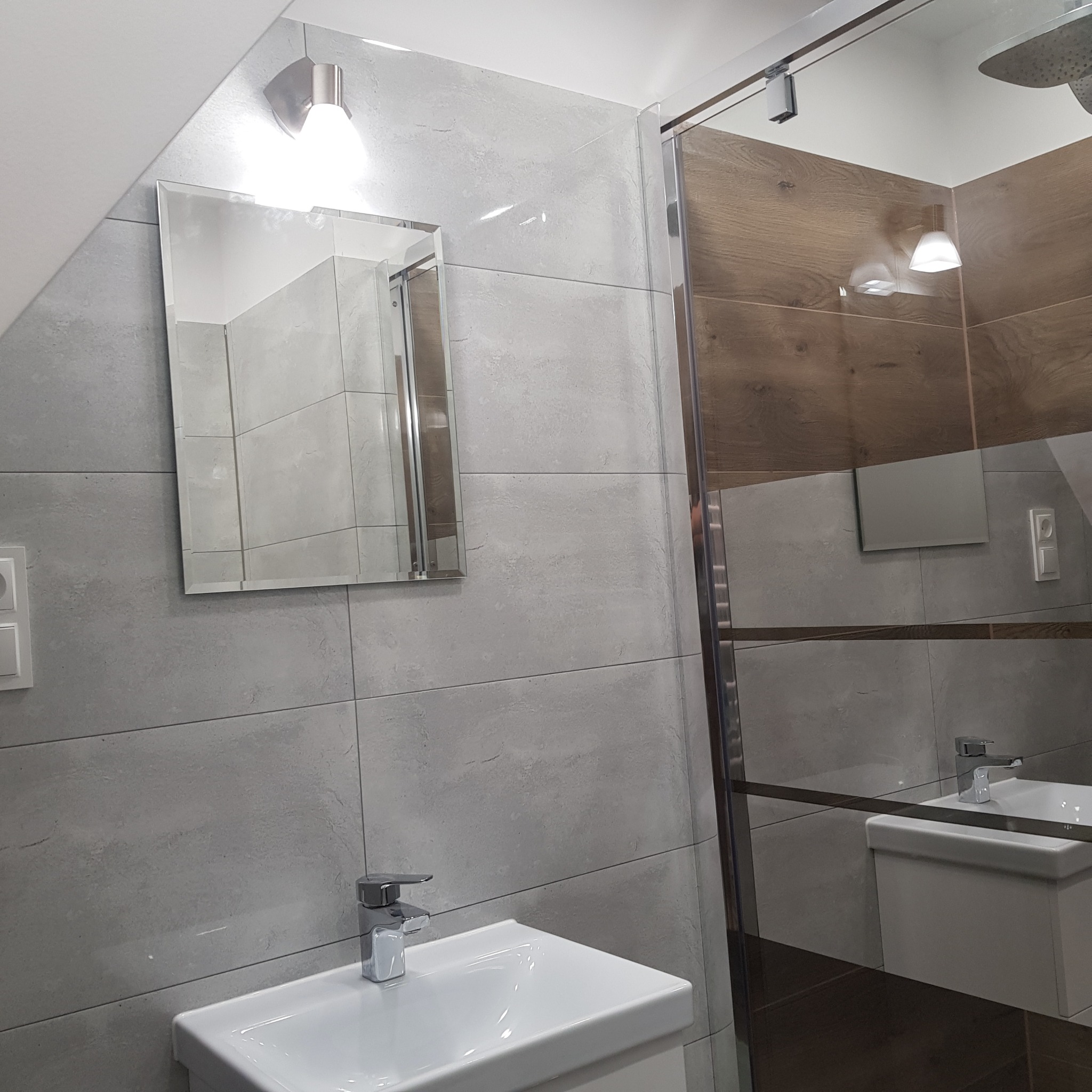 Nowoczesna łazienka z szarymi płytkami, lustrem z oświetleniem i kabiną prysznicową z drewnopodobnymi panelami na górze, widok na umywalkę z chromowaną baterią.
