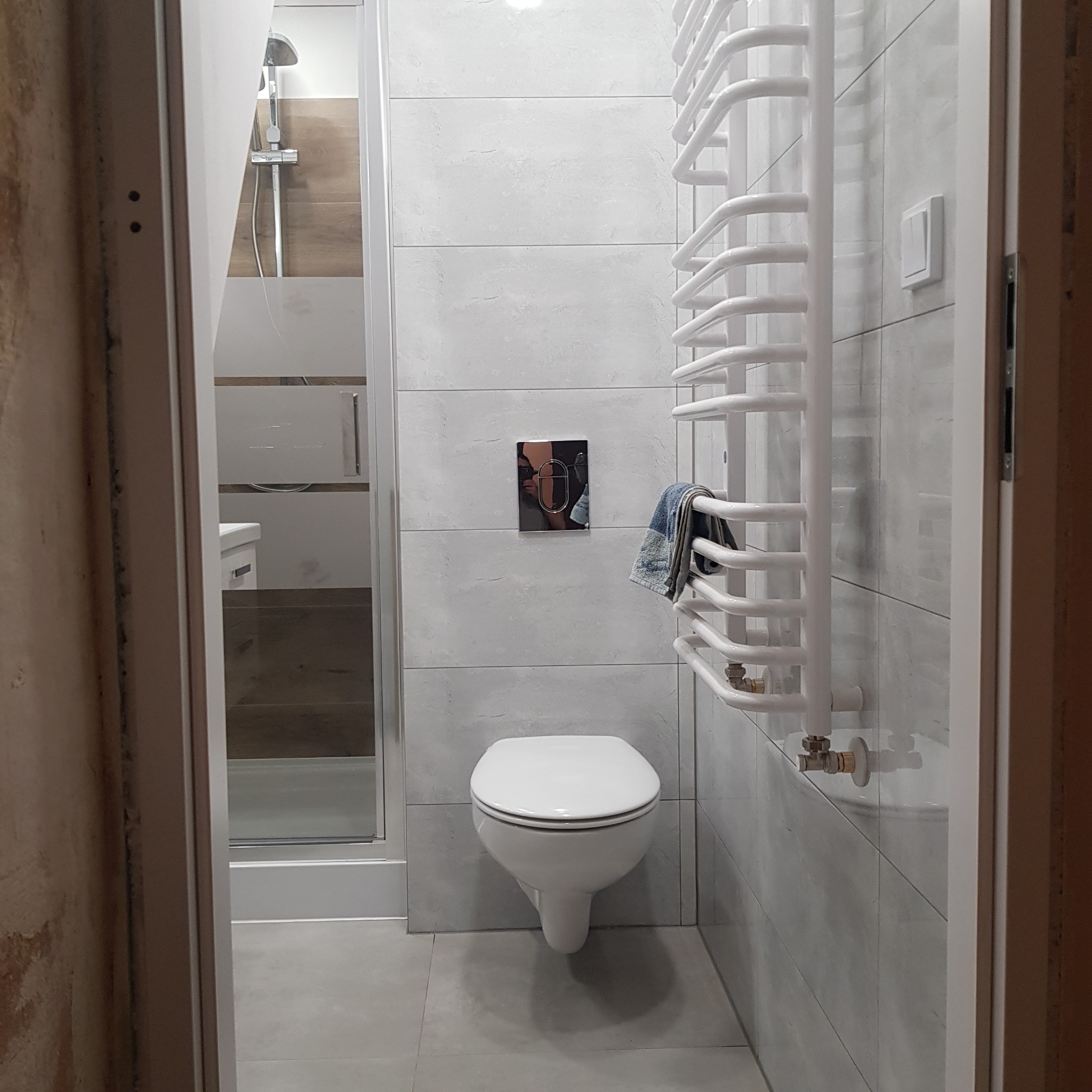 Nowoczesna łazienka z szarymi płytkami, wiszącą toaletą, grzejnikiem drabinkowym i kabiną prysznicową z widoczną słuchawką. Na grzejniku powieszony jest ręcznik w odcieniach niebieskiego i szarego.
