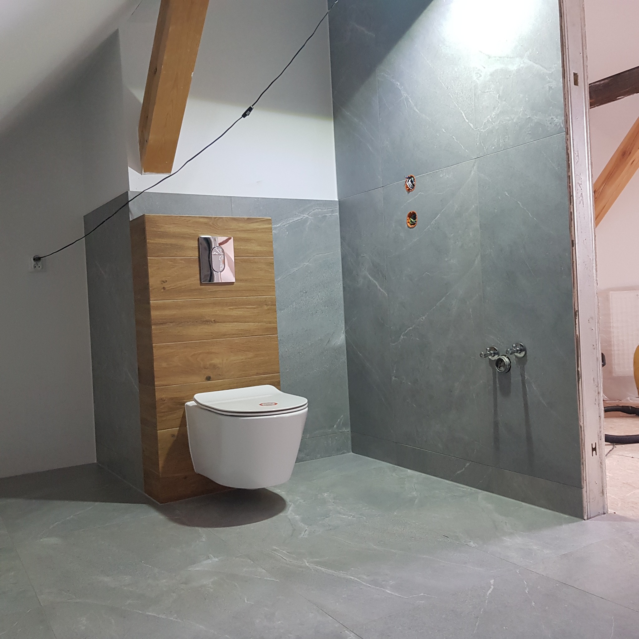 Wisząca toaleta z drewnianą obudową i chromowanym przyciskiem spłukiwania na tle szarych płytek imitujących marmur w łazience na poddaszu z widoczną drewnianą belką stropową.