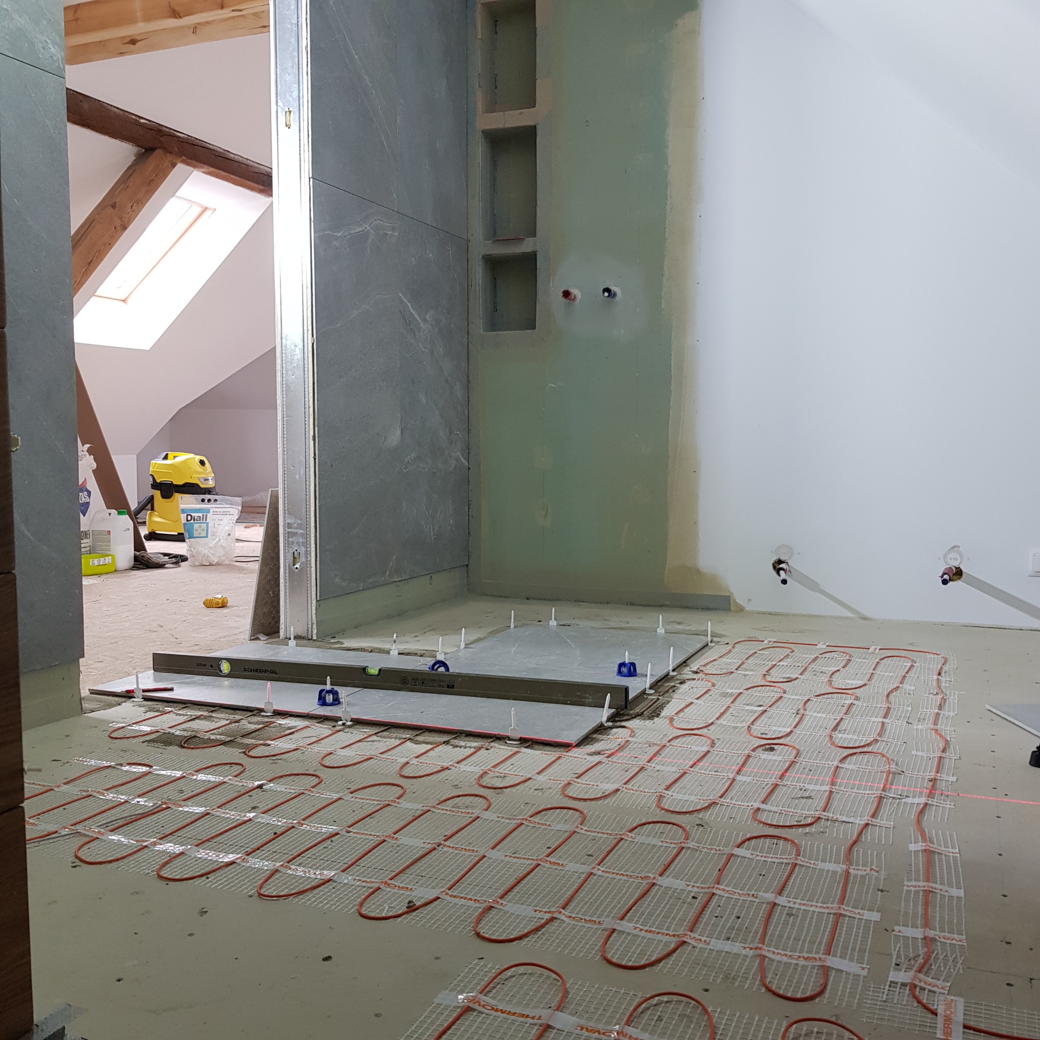 Montaż ogrzewania podłogowego w łazience na poddaszu, widoczne płytki, poziomica, krzyżyki dystansowe, rury grzewcze Thermoval oraz przygotowane ściany z wnękami.