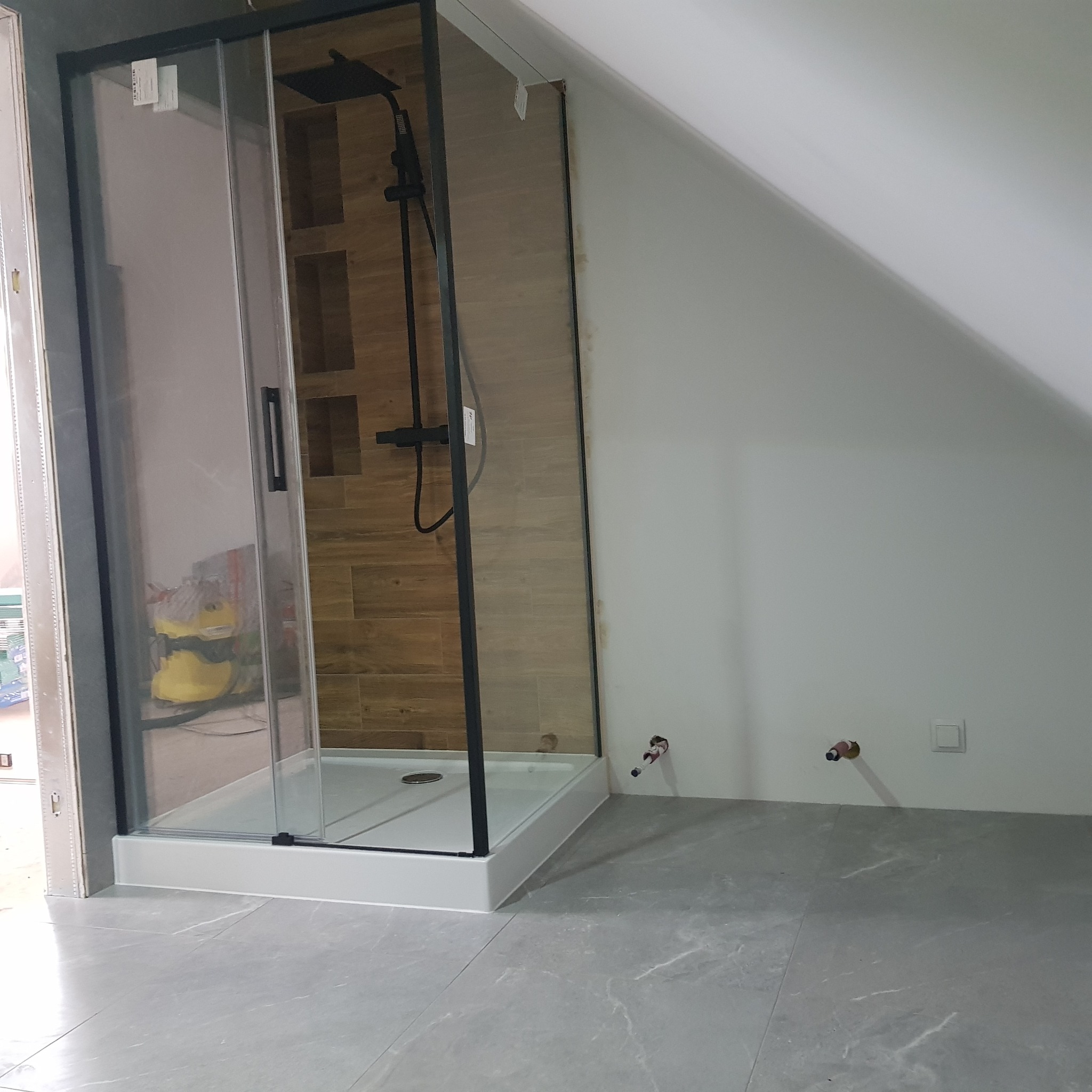 Nowa kabina prysznicowa z czarnymi profilami zamontowana w łazience na poddaszu, widoczne przyłącza wodne i fragment ściany z drewnopodobnymi płytkami.