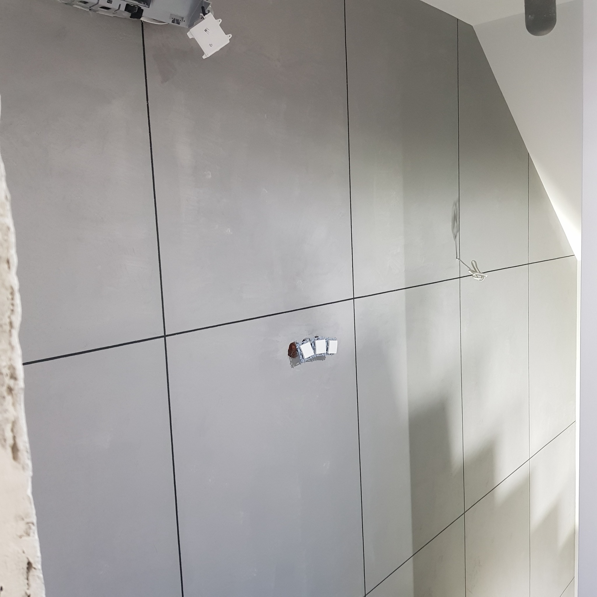 Ściana pokryta szarymi panelami z widocznymi fugami, otwory na gniazdka elektryczne w trakcie przygotowania, widoczne kable.