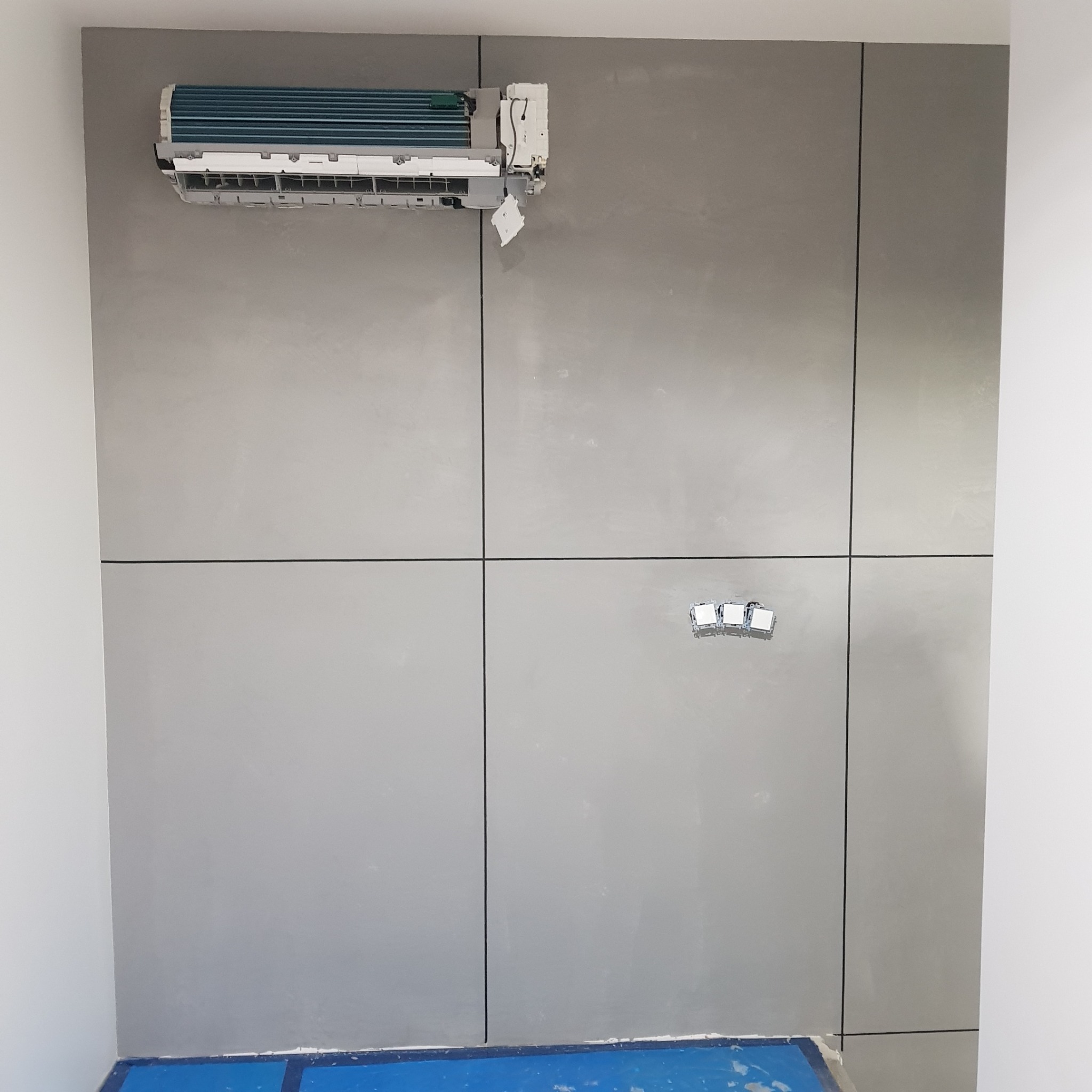 Ściana z szarych paneli z widocznymi spoinami, zamontowany klimatyzator i puszki elektryczne, zabezpieczona podłoga.