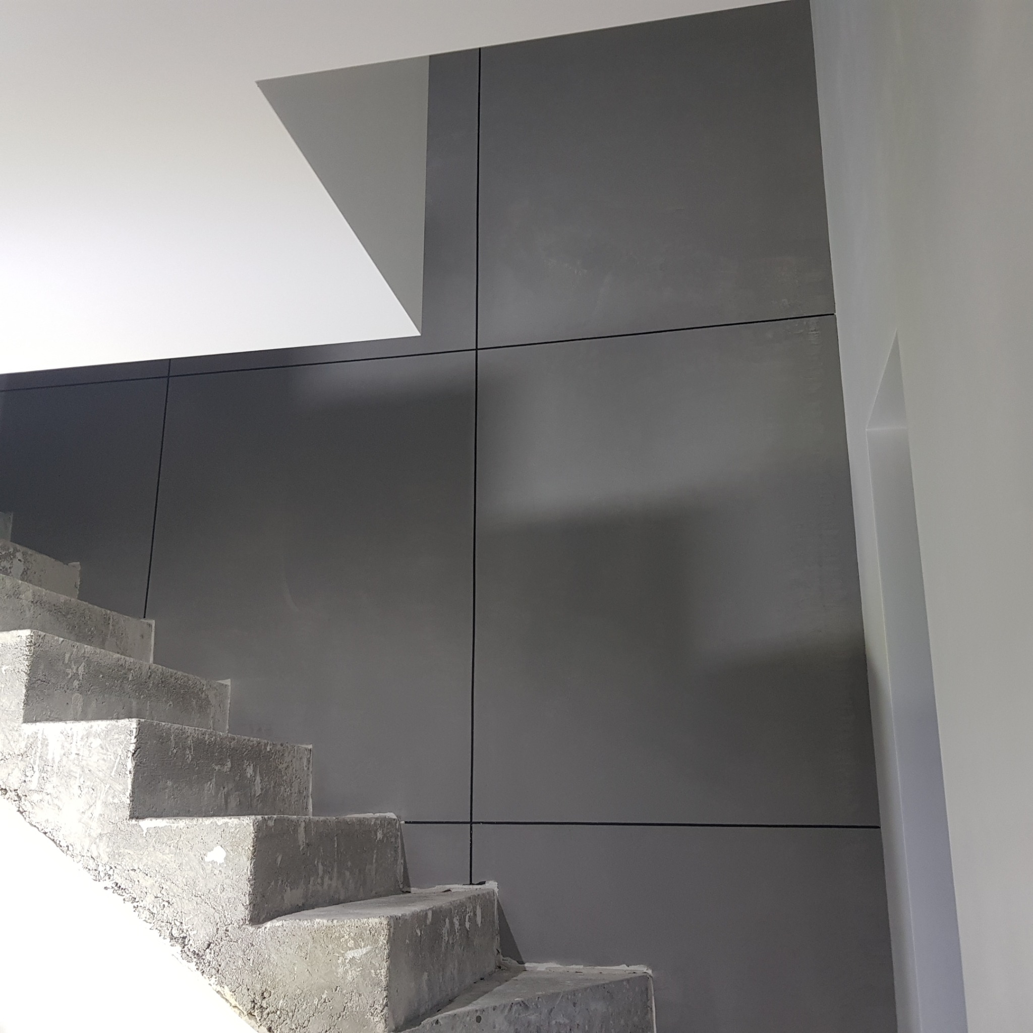 Surowe, betonowe schody prowadzące w górę, na tle szarej ściany z panelami oddzielonymi czarnymi liniami, widoczne przygotowania do dalszych prac wykończeniowych.
