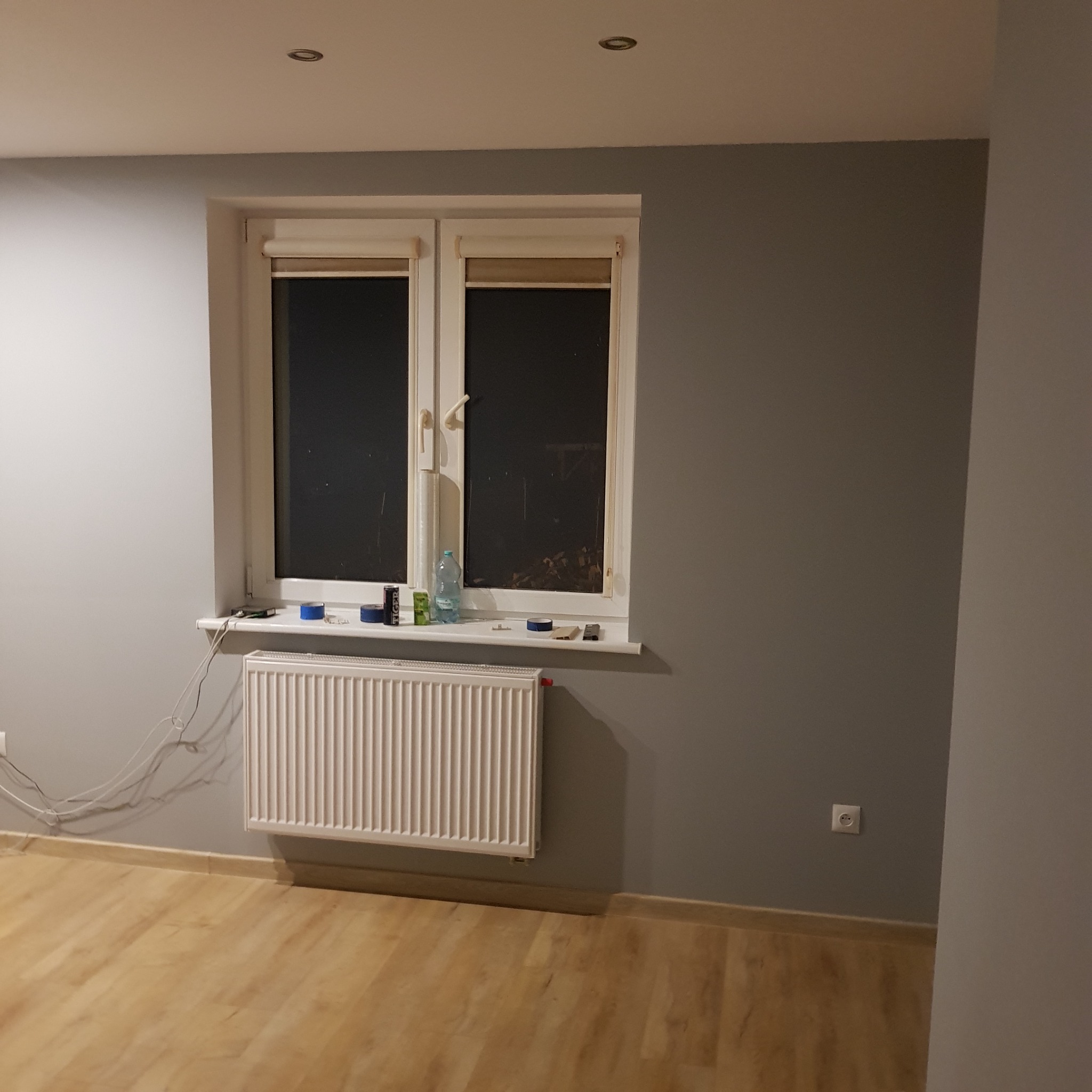 Pokój po malowaniu ścian na szaro, widoczny grzejnik, okno z roletami i przedmioty na parapecie.