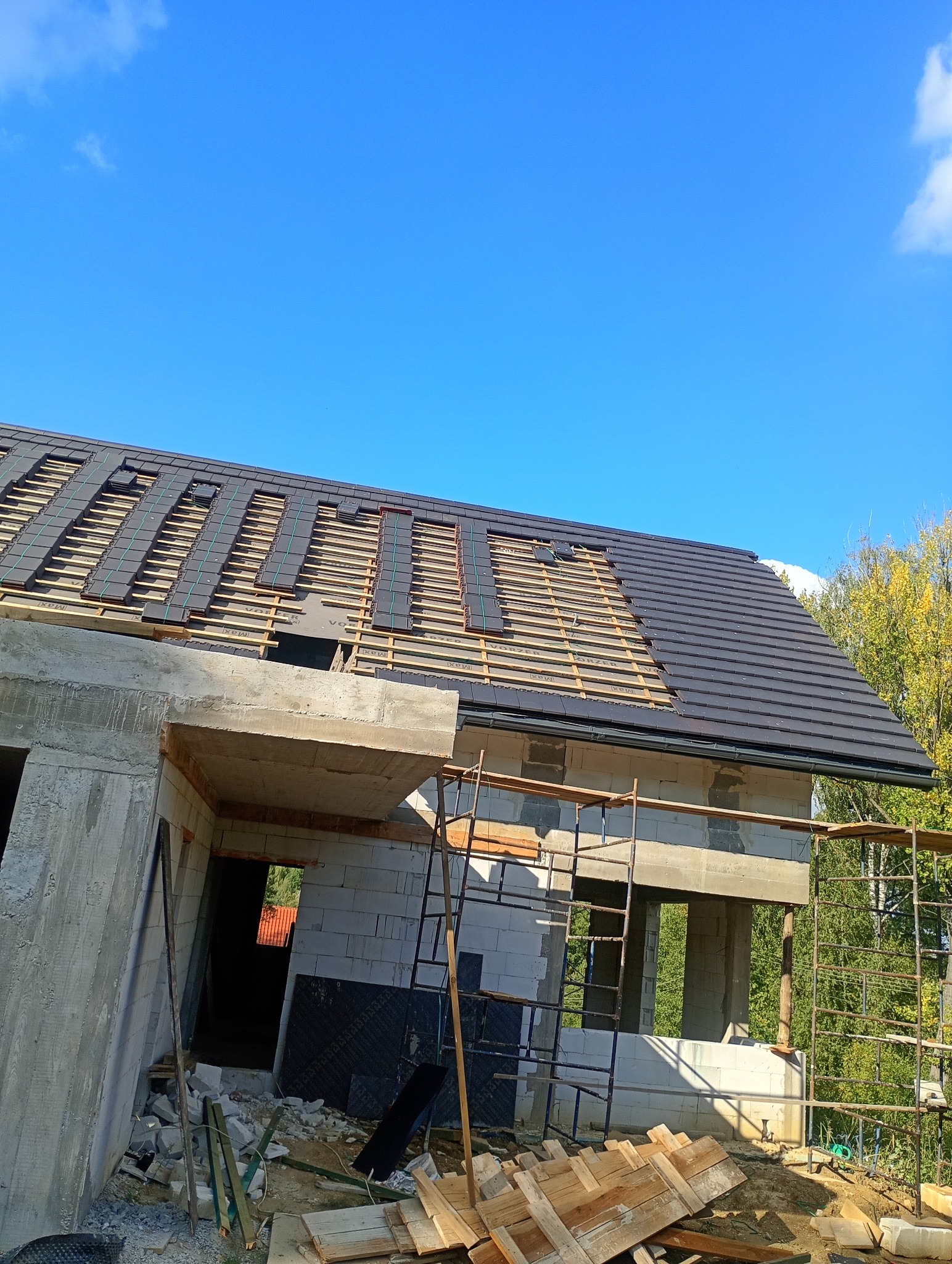 Budowa domu: fragment dachu z częściowo ułożoną, ciemną dachówką ceramiczną na łatach, widoczne ściany z betonu komórkowego i rusztowania. Błękitne niebo w tle.