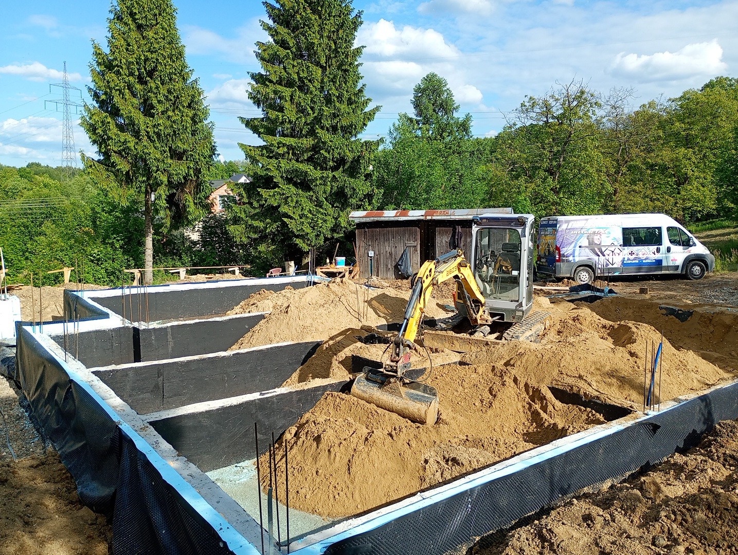 Fundamenty domu z hydroizolacją i mini koparką w środku wykopu. W tle zieleń drzew i bus firmy budowlanej. Etap prac ziemnych przed wylaniem płyty fundamentowej.