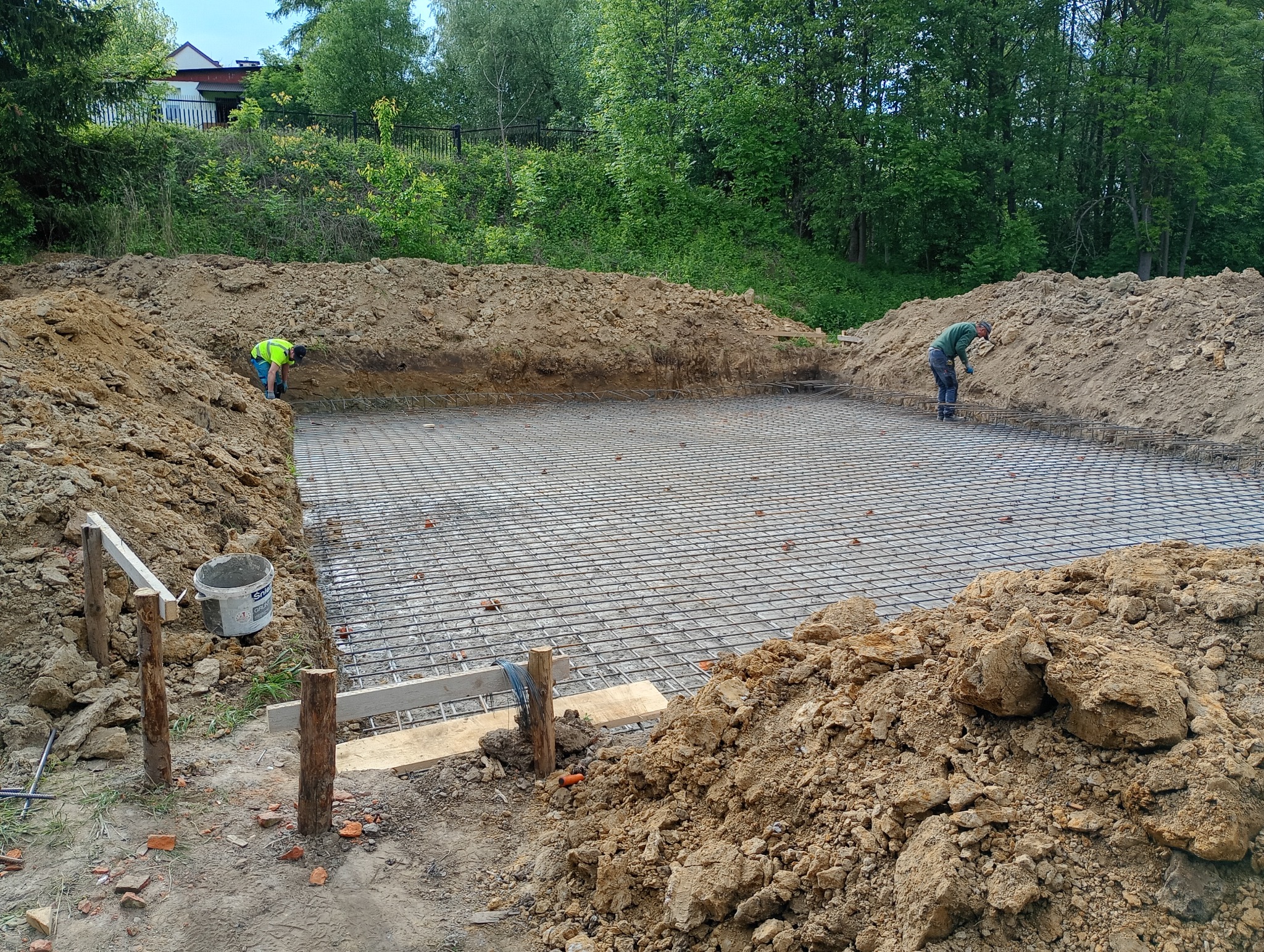 Zbrojenie fundamentów pod budowę domu jednorodzinnego. Dwóch pracowników w trakcie prac ziemnych i układania siatki zbrojeniowej. Widoczne narzędzia i materiały budowlane.