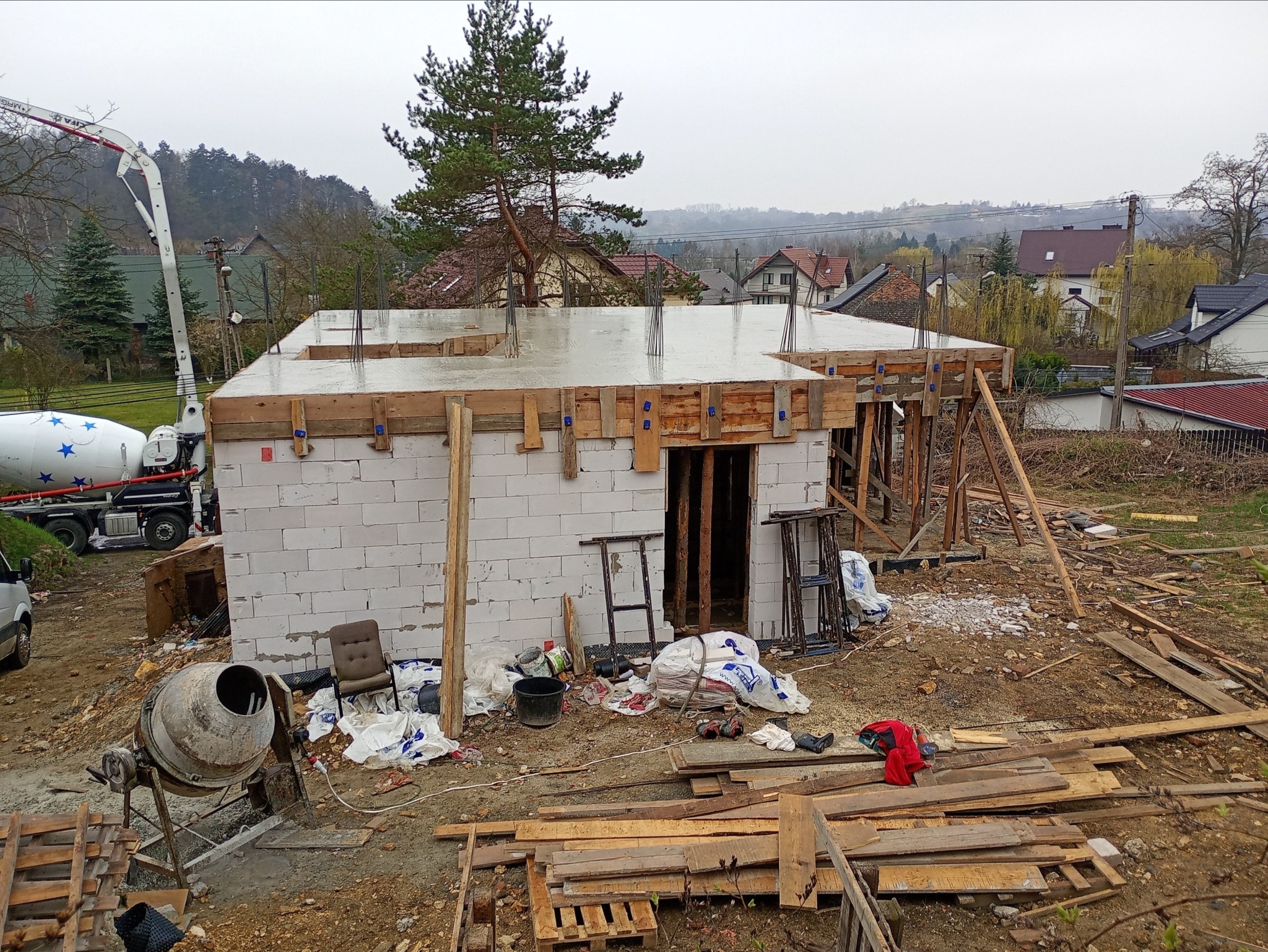 Budowa domu w trakcie: wylany strop na ścianach z betonu komórkowego, widoczny mikser do betonu i rozgardiasz na placu budowy. Ujęcie z zewnątrz, dzień.
