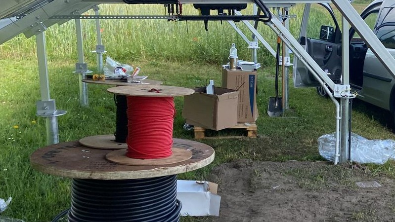 Szkielet konstrukcji pod panele fotowoltaiczne na łące, z widocznymi bębnami z przewodami elektrycznymi: czerwonym, czarnym i grubym czarnym kablem, obok otwartego samochodu i narzędzi.