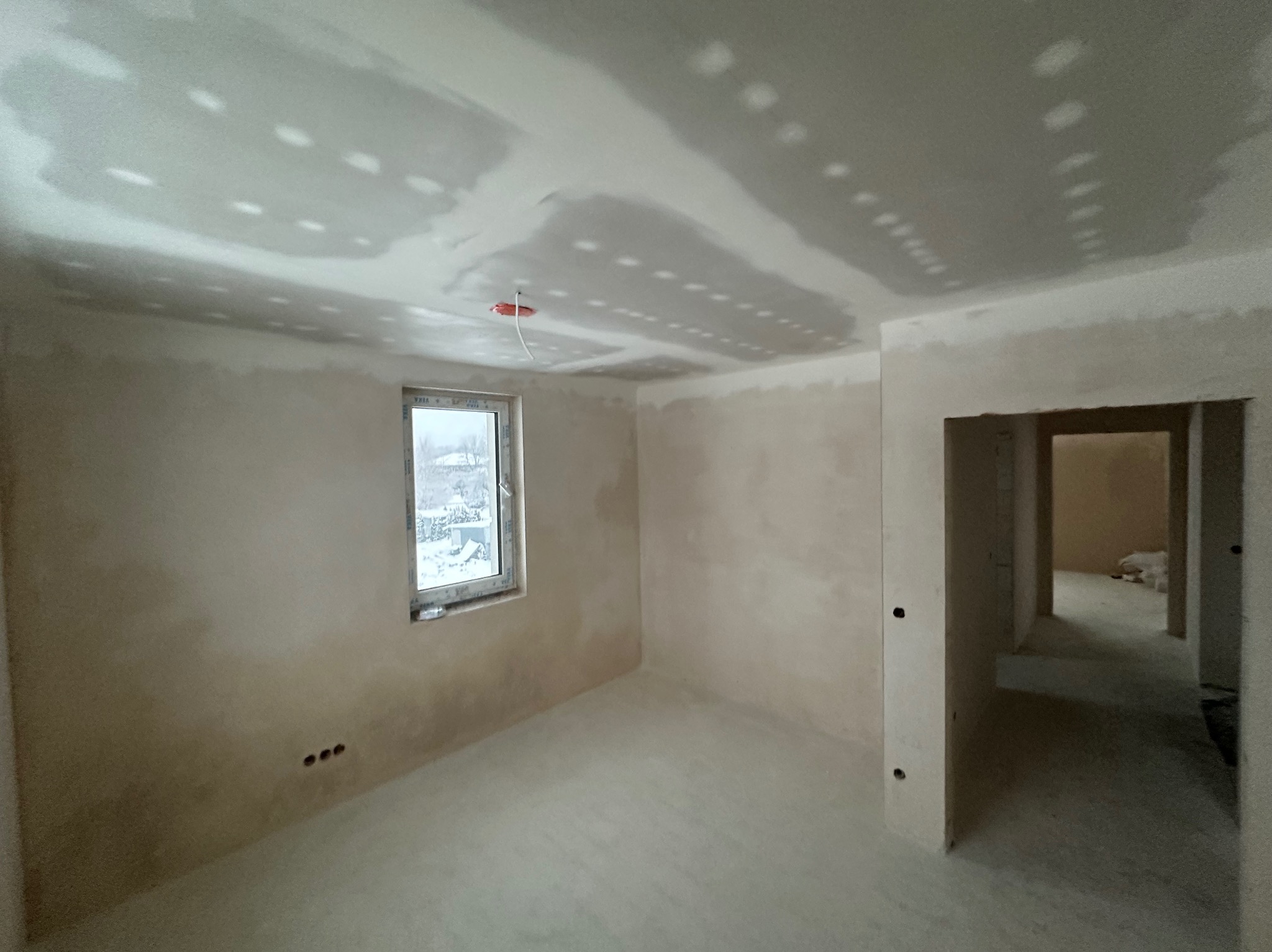 Wnętrze pokoju w trakcie prac wykończeniowych. Widoczne ściany po szpachlowaniu, sufit z widocznymi punktami mocowania, okno z widokiem na zewnątrz i otwór drzwiowy.