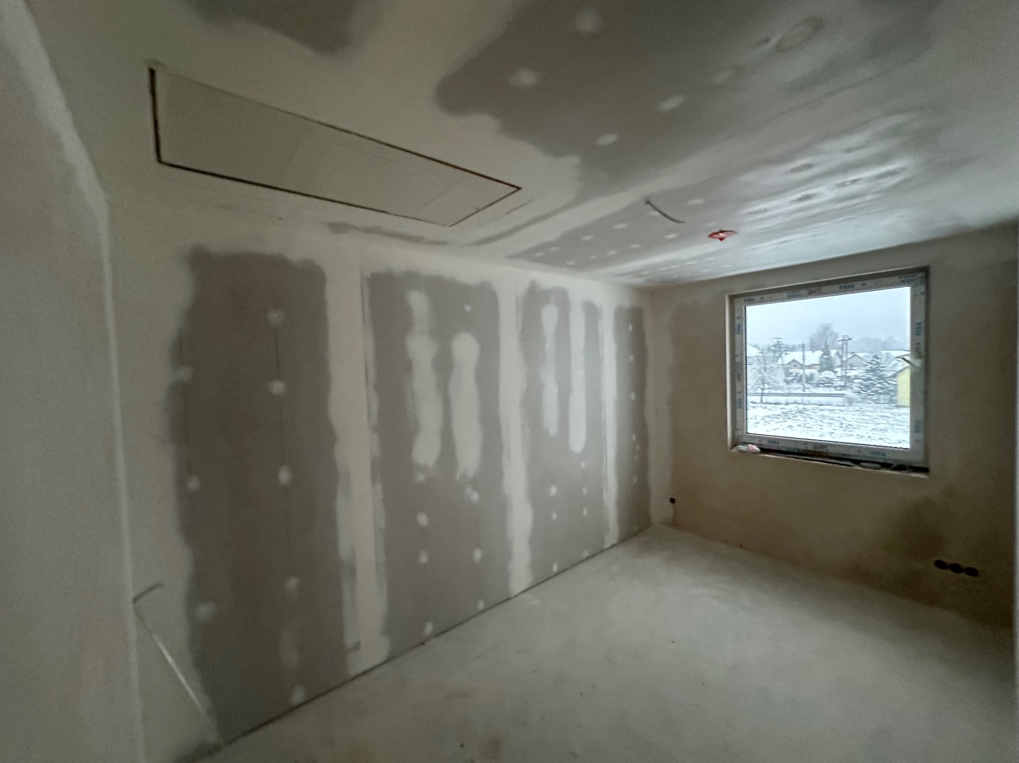 Pokój w trakcie prac wykończeniowych: ściany z widocznymi łączeniami płyt, okno z widokiem na zimowy krajobraz, sufit z otworem rewizyjnym.
