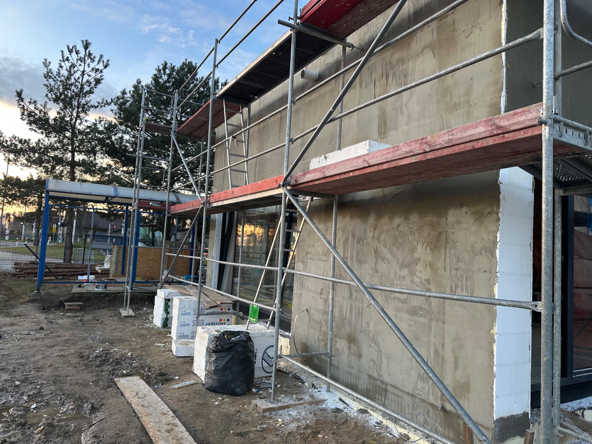 Ściana budynku w trakcie ocieplania, widoczne rusztowania, styropian i częściowo nałożony tynk. Ujęcie z dołu, w tle drzewa i błękitne niebo.