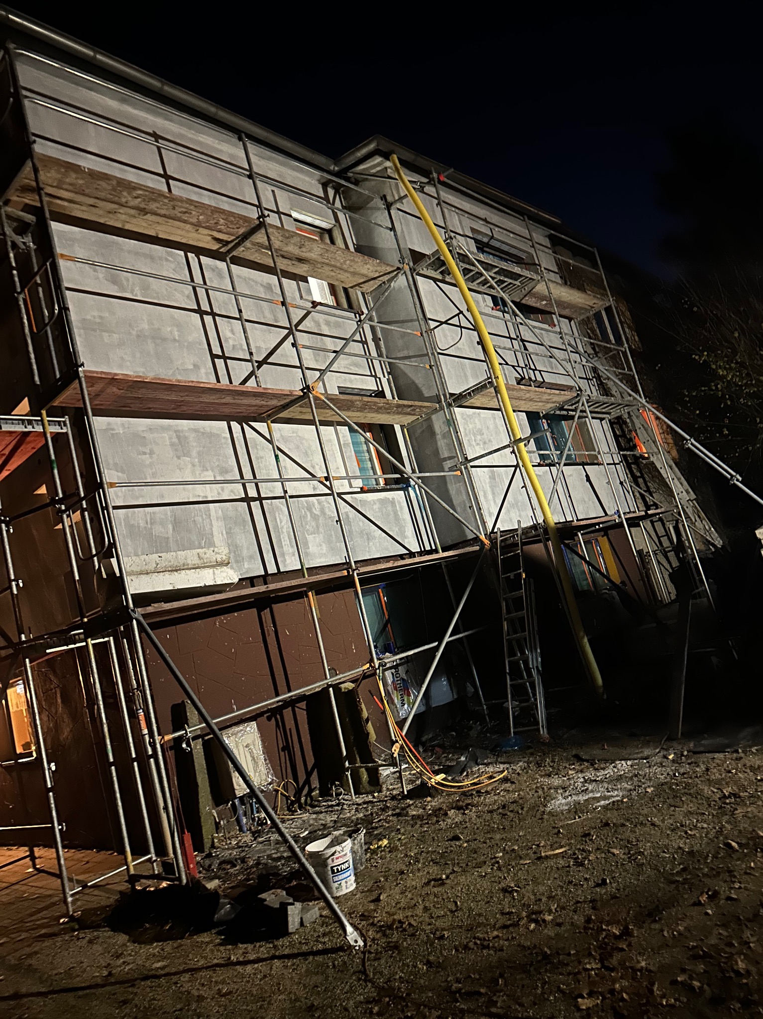 Budynek w trakcie tynkowania, otoczony rusztowaniami. Widoczny kubeł z tynkiem i oświetlenie nocne. Prace na zewnątrz budynku.