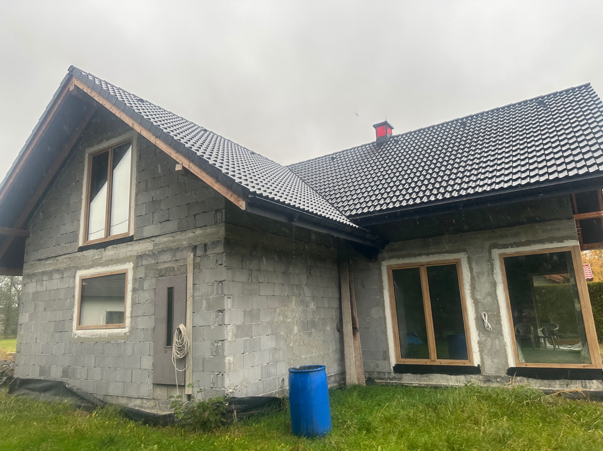 Surowy stan domu z betonu komórkowego z oknami w drewnianych ramach i czarnym dachem. Widoczna niebieska beczka i lina. Etap budowy przed tynkowaniem.