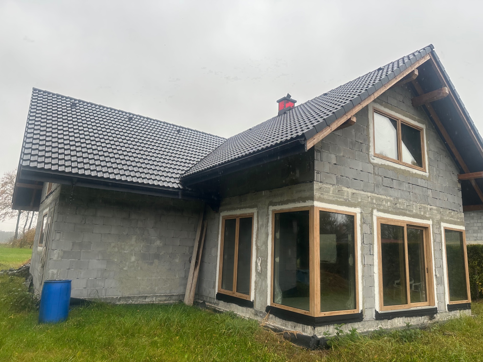 Surowy dom z szarego bloczka z zamontowanymi oknami z drewnianymi ramami, czarnym dachem i czerwonym kominem, otoczony zieloną trawą i błękitną beczką.