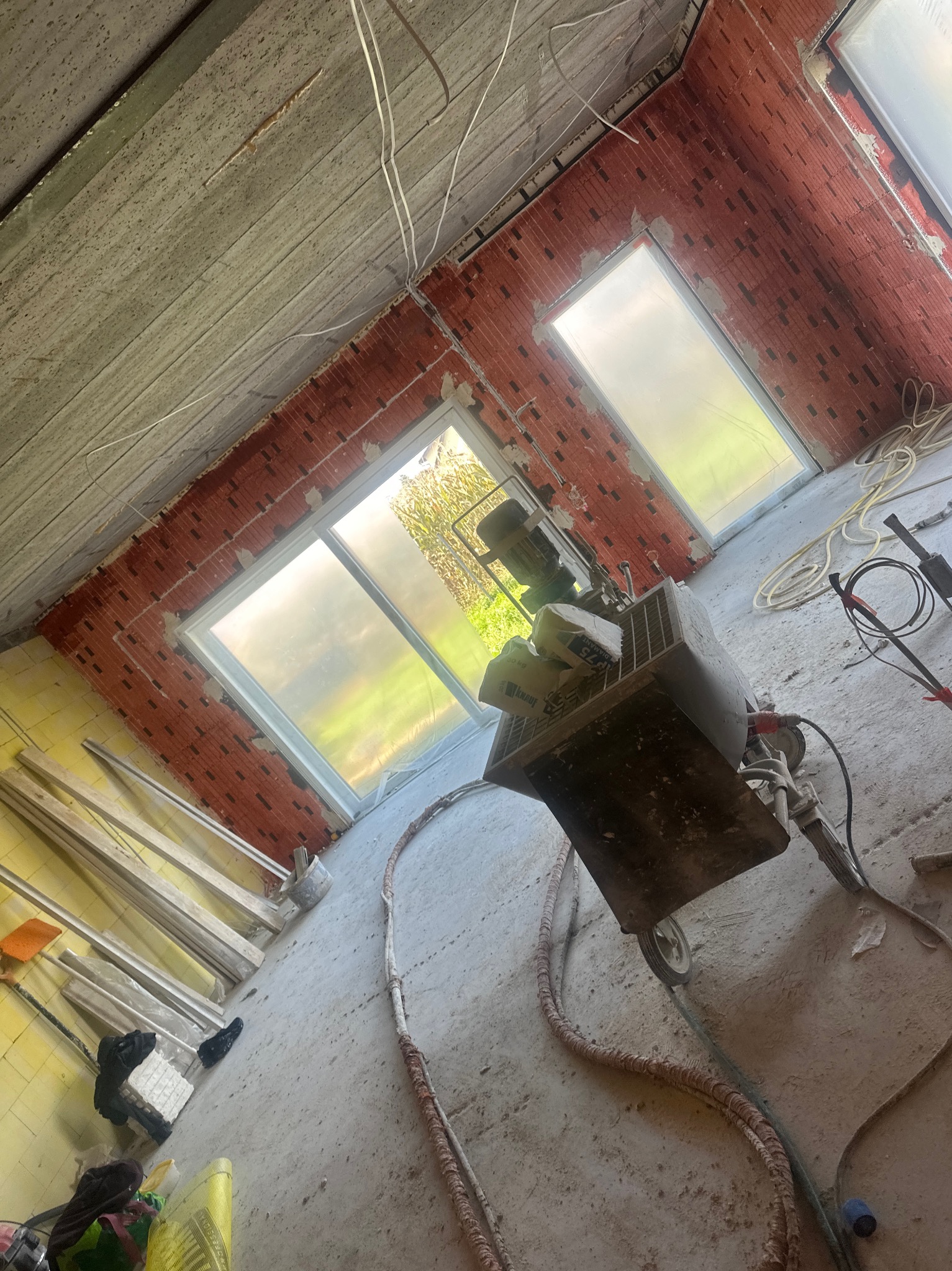 Wnętrze w trakcie prac: ściany z cegły, okna zabezpieczone folią, maszyna do tynkowania z workami zaprawy. Widoczne przewody i narzędzia. Surowy stan wykończenia.