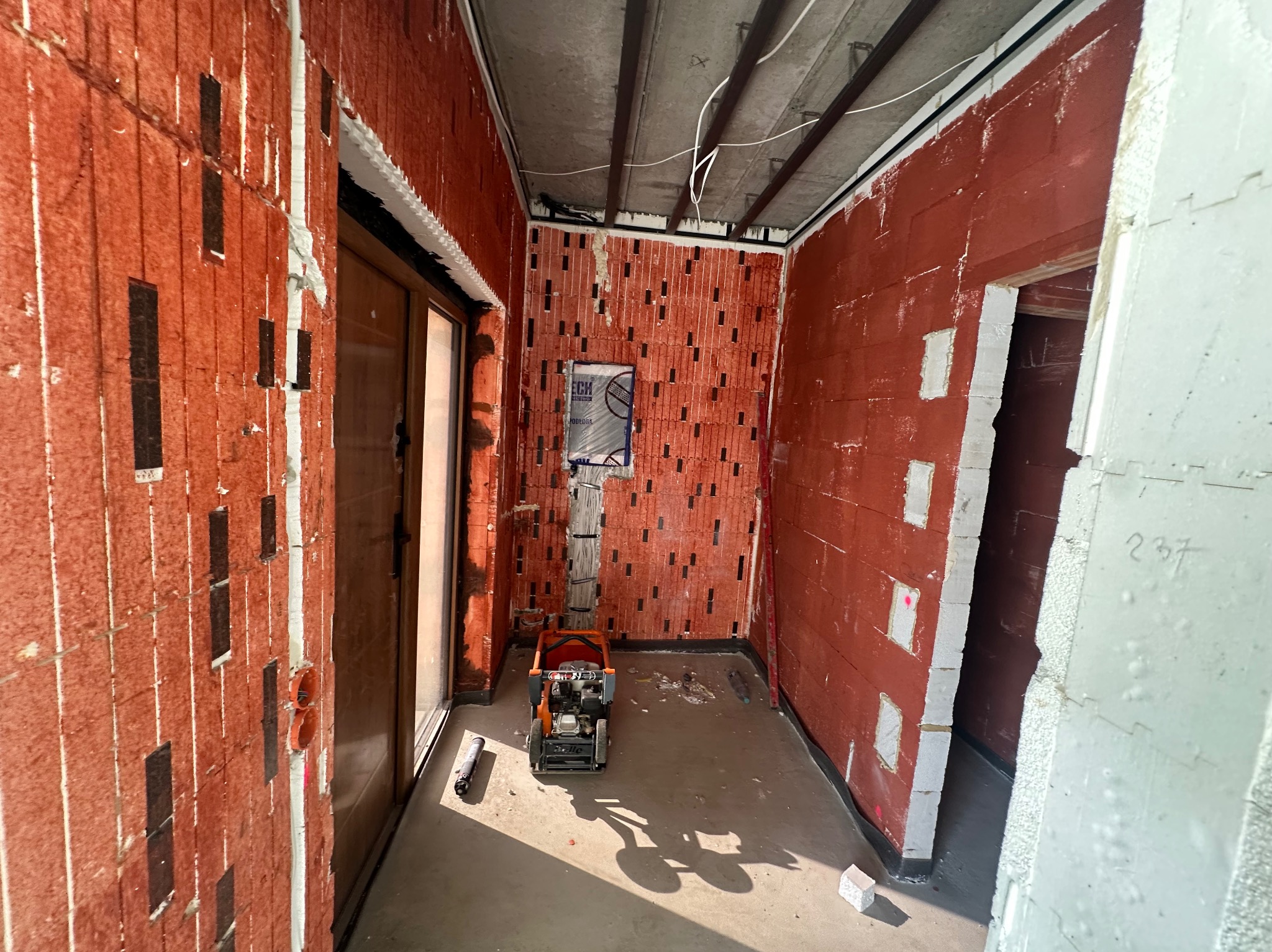 Wnętrze w trakcie prac wykończeniowych: czerwone ściany z pustaków, częściowo ocieplone, z widoczną maszyną do zagęszczania podłoża i instalacjami elektrycznymi.