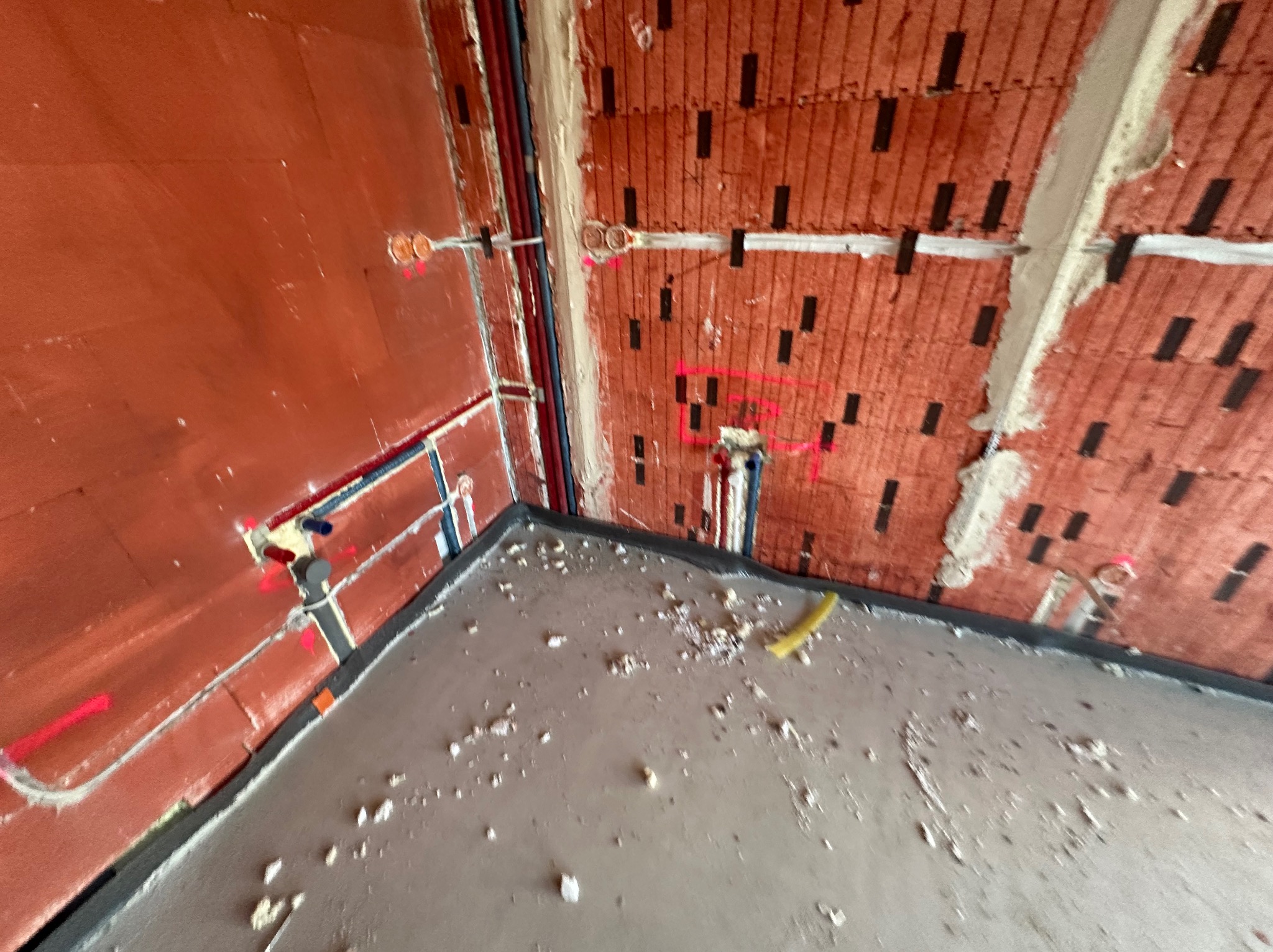 Surowy stan wnętrza: ściany z czerwonej cegły z instalacjami elektrycznymi i hydraulicznymi, wylewka podłogowa z widocznymi zabrudzeniami, przygotowane do dalszych prac.