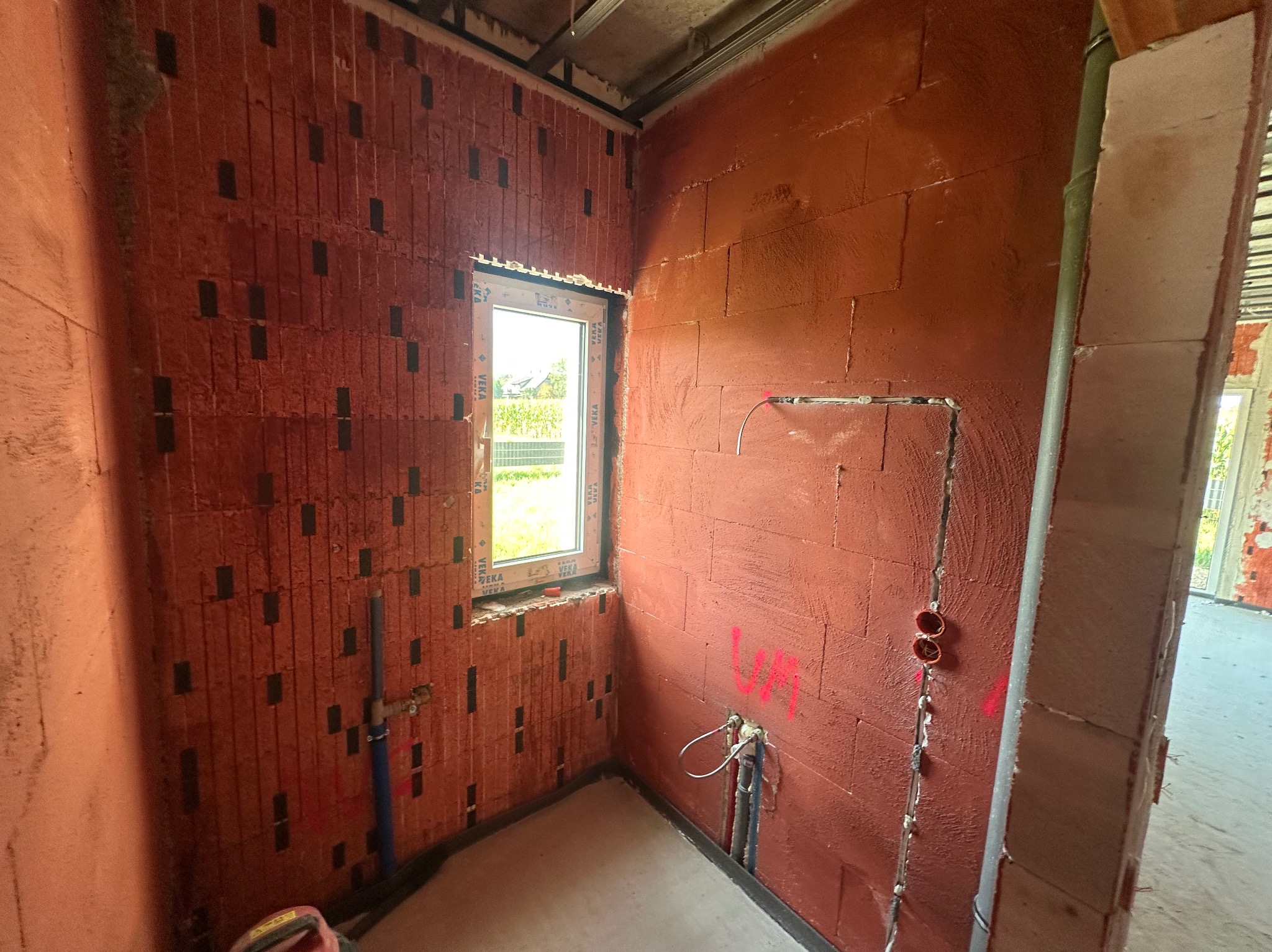 Surowy stan wnętrza: ściany z czerwonej cegły z widocznymi instalacjami wodnymi i elektrycznymi. Okno z widokiem na zieleń. Podłoga betonowa. W tle widoczne kolejne pomieszczenie.