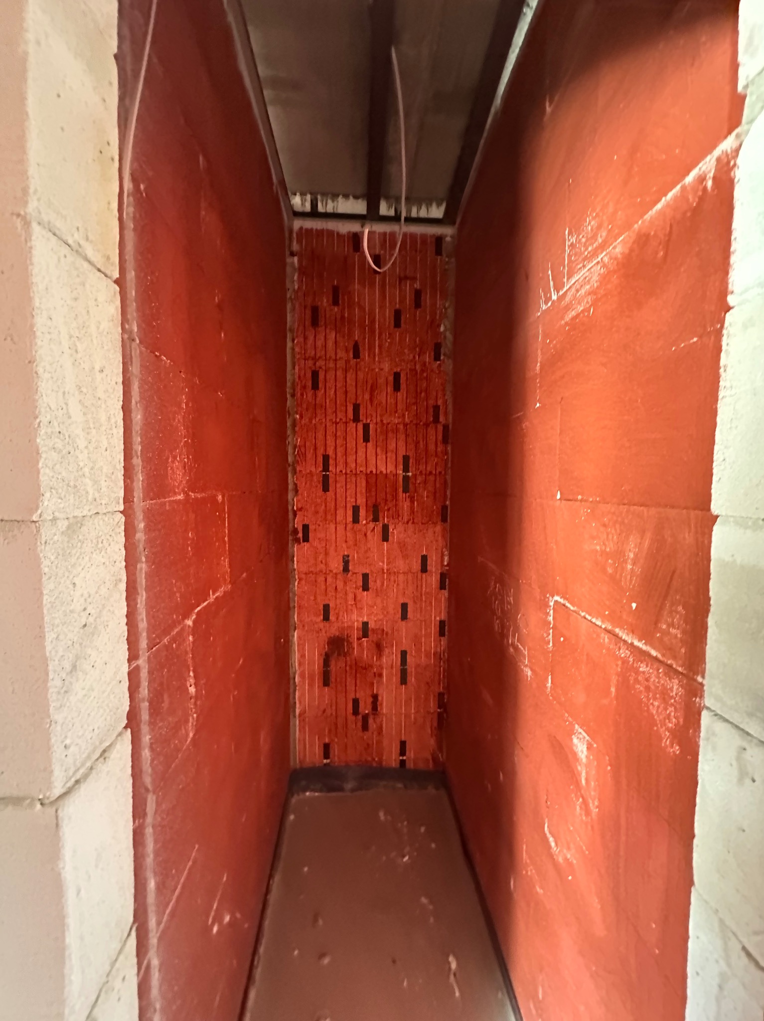 Wnętrze w trakcie prac wykończeniowych: ściany pokryte czerwoną warstwą izolacyjną, widoczne białe narożniki i fragment sufitu z instalacją.