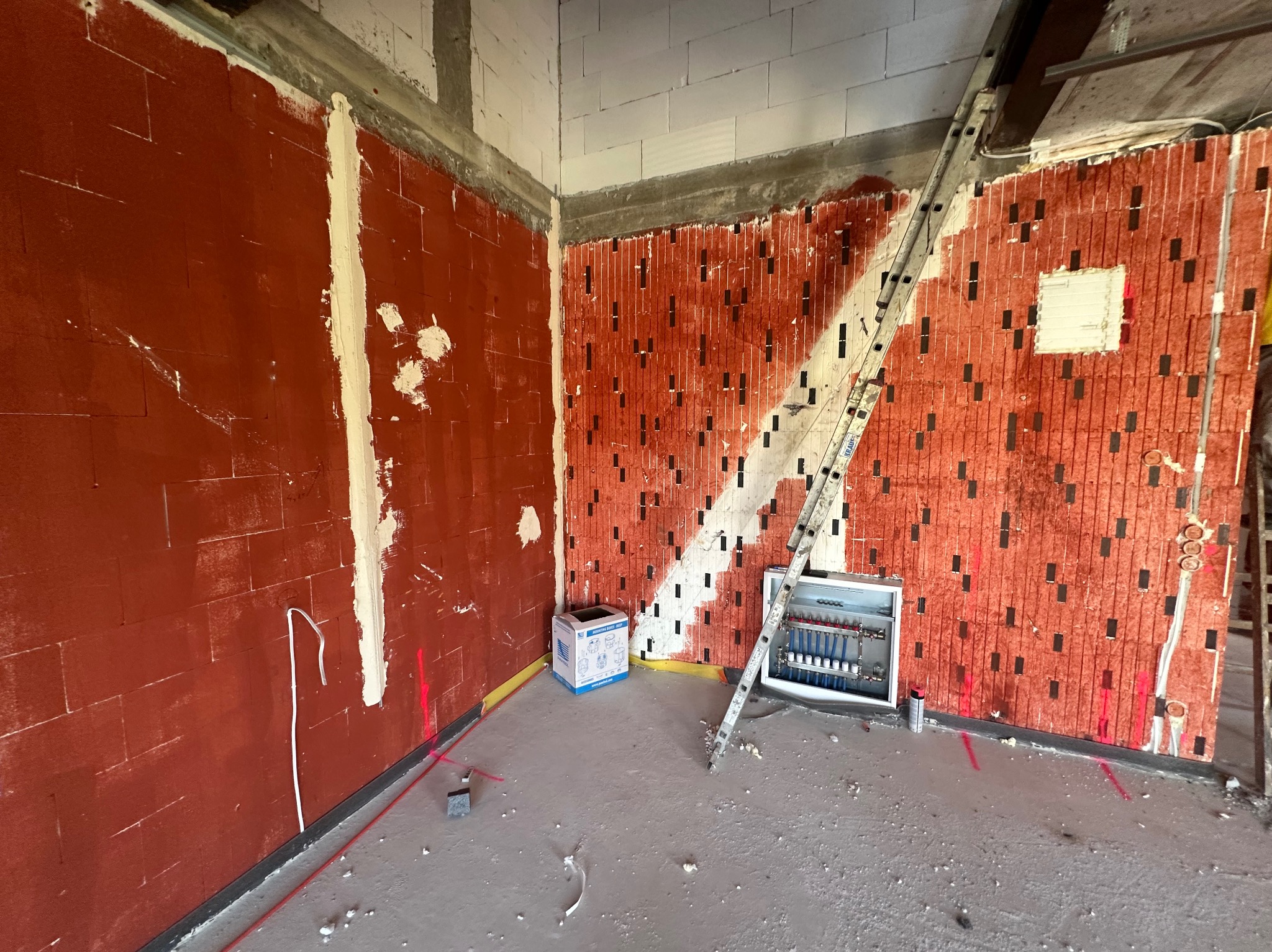Czerwone ściany z pustaków ceramicznych z pionowymi nacięciami, częściowo pokryte białą zaprawą. Widoczna drabina i rozdzielacz ogrzewania podłogowego. Stan surowy.