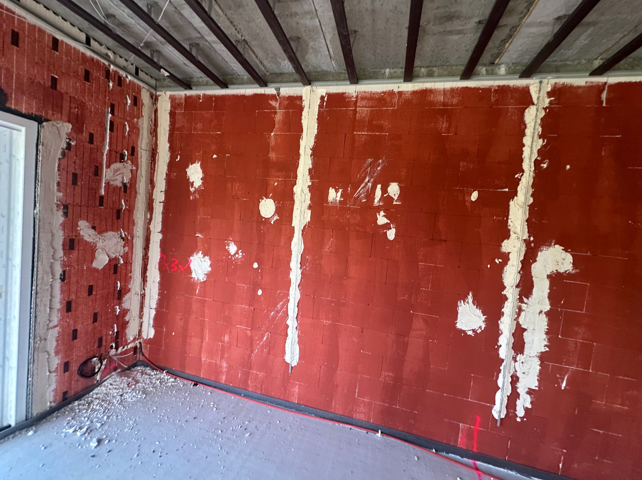 Czerwona ściana z cegły ceramicznej w trakcie prac przygotowawczych, z widocznymi śladami po szpachlowaniu i instalacjach elektrycznych. Surowy betonowy sufit z metalowymi profilami.
